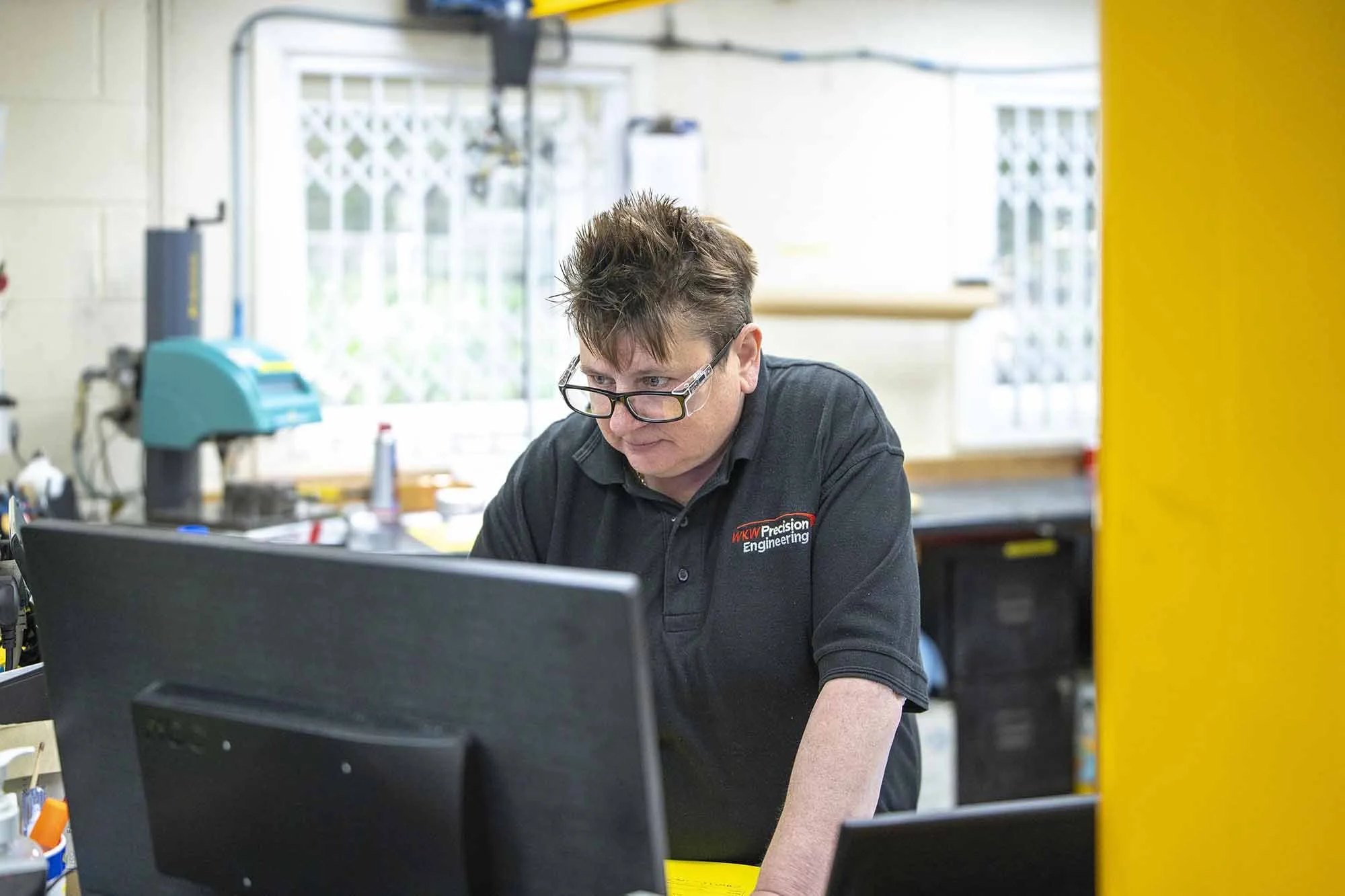 A man working at a desk in a workshop or industrial setting, wearing a black polo shirt with a logo that reads 'WKW Precision Engineering,' glasses, and focused on a computer monitor.