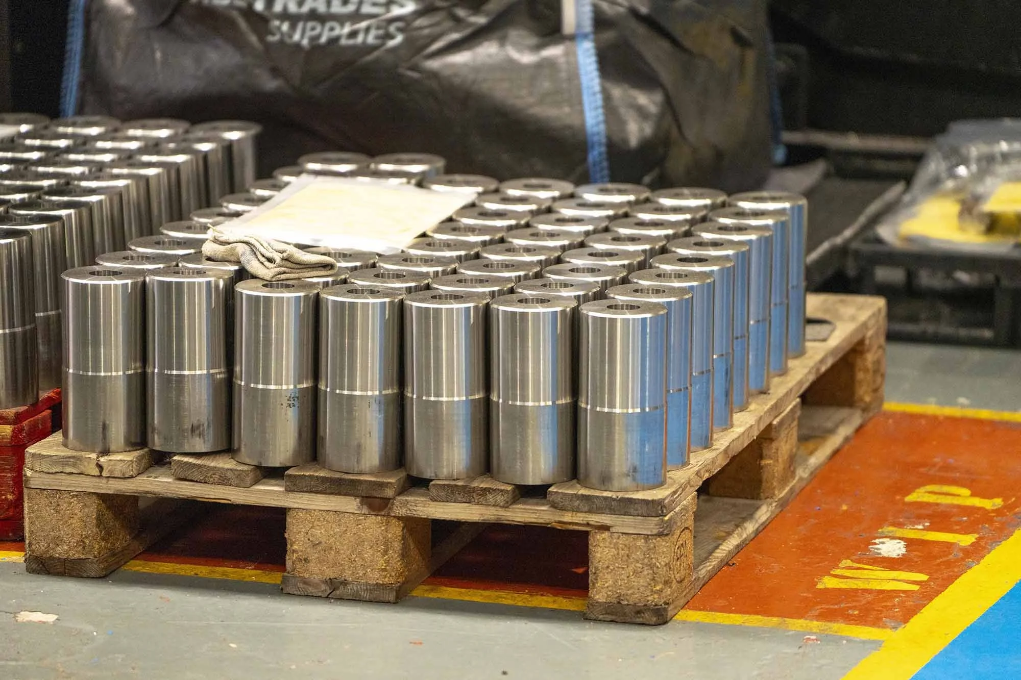 Multiple metal cylinders arranged on a wooden pallet, with a rag and a paper on top, in an industrial or warehouse setting.