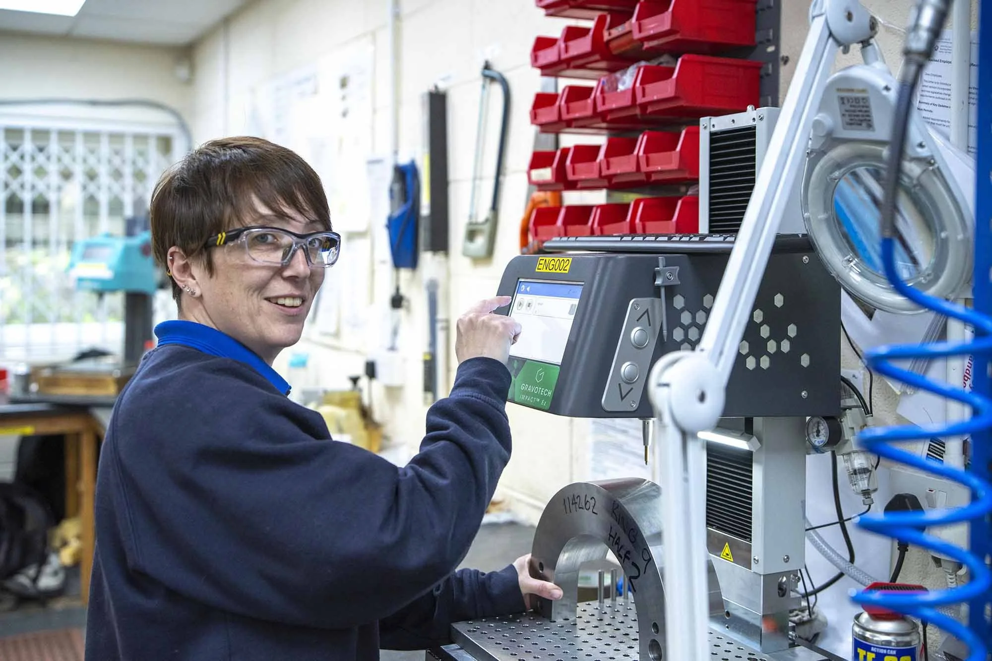 A woman wearing safety glasses in a workshop, operating a digital control panel on a machine with a metal component.