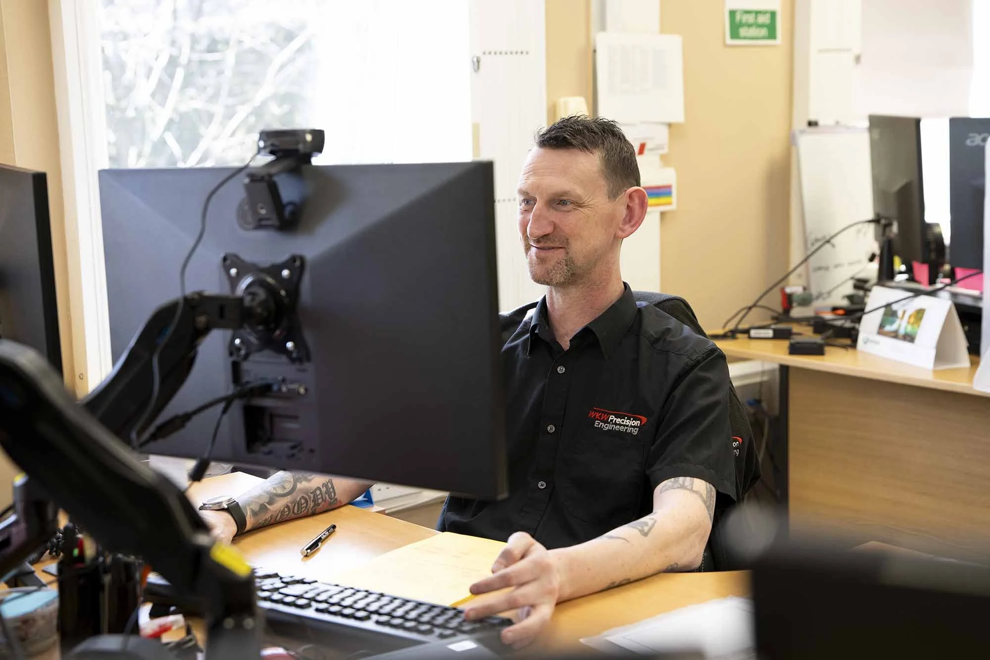 A man with tattoos wearing a black shirt with 'WKY Precision Engineering' logo, working at a computer in an office.