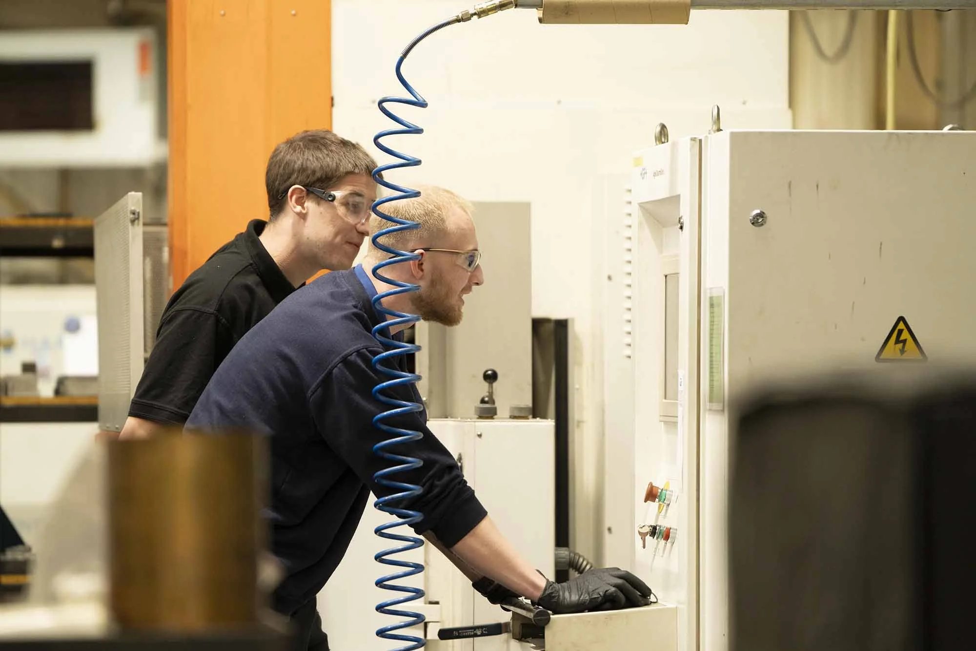 Two men working in a laboratory, wearing safety goggles and black gloves, operating a machine with control panels and safety warning signs.