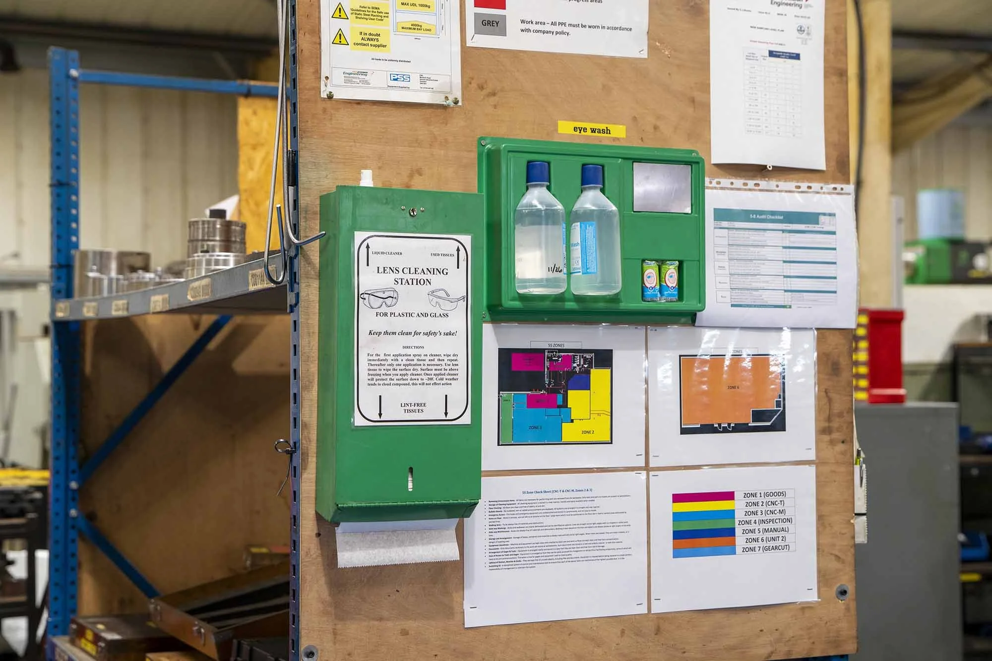 A safety station with a green lens cleaning box, spray bottles, and eye wash station on a wood panel in a warehouse or industrial setting, with various safety and layout maps posted on the wall.