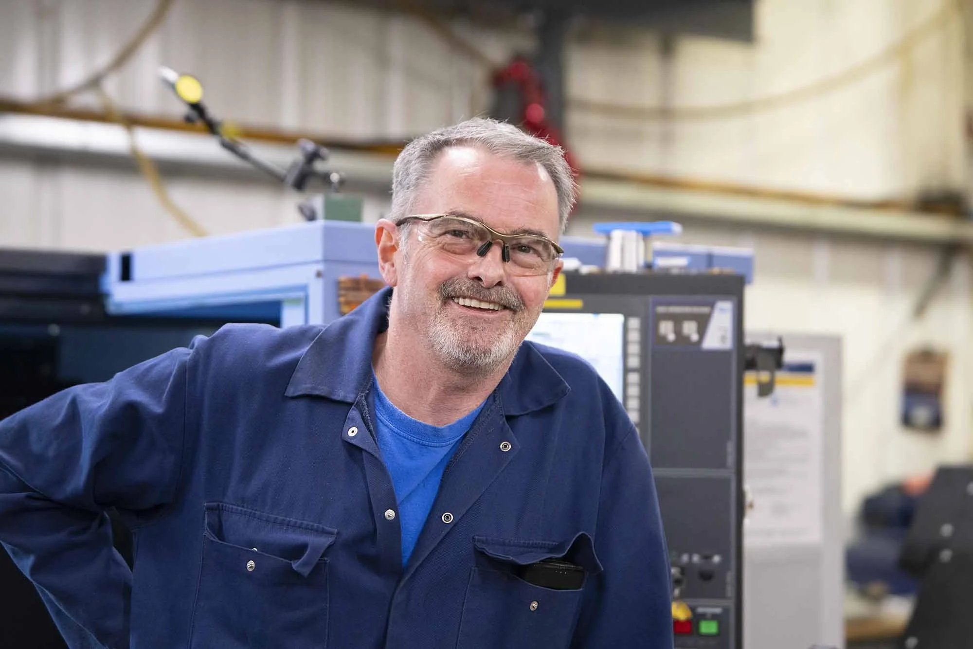 A smiling middle-aged man with glasses, sporting a gray beard, wearing a blue work jacket, standing in an industrial or workshop setting with machinery in the background.