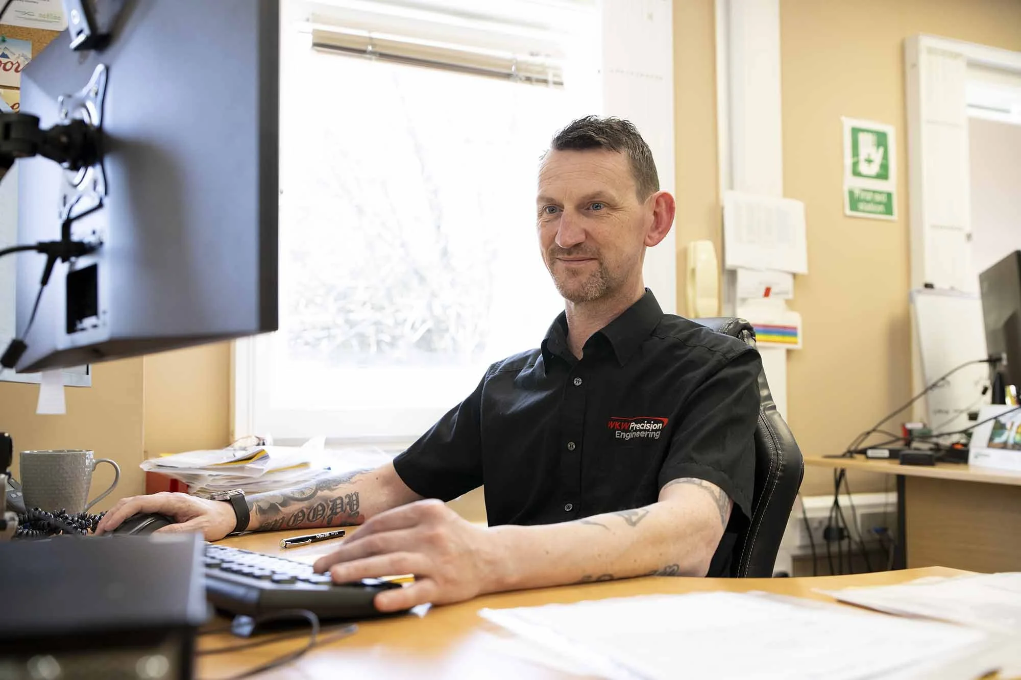 A man sitting at a desk in an office, working on a computer, wearing a black shirt with a company logo, with papers and a coffee mug on the desk.