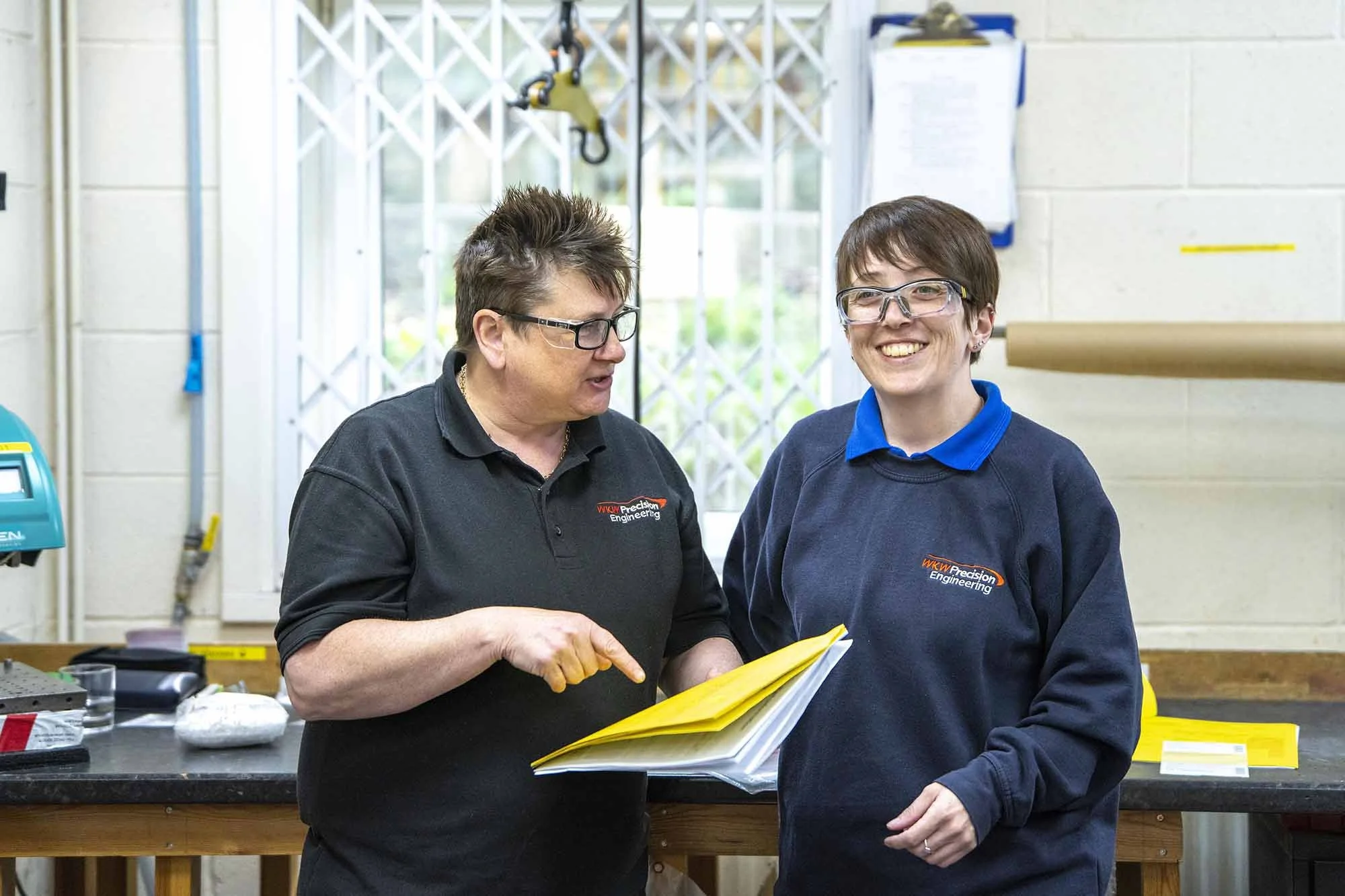 Two women in safety glasses and dark work shirts, smiling and discussing documents in a workshop or industrial setting.