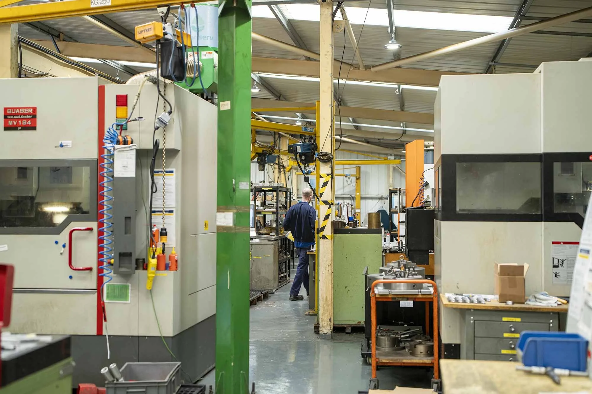 Industrial manufacturing workshop with machinery, tools, and a worker in safety clothing, surrounded by equipment and workstations.
