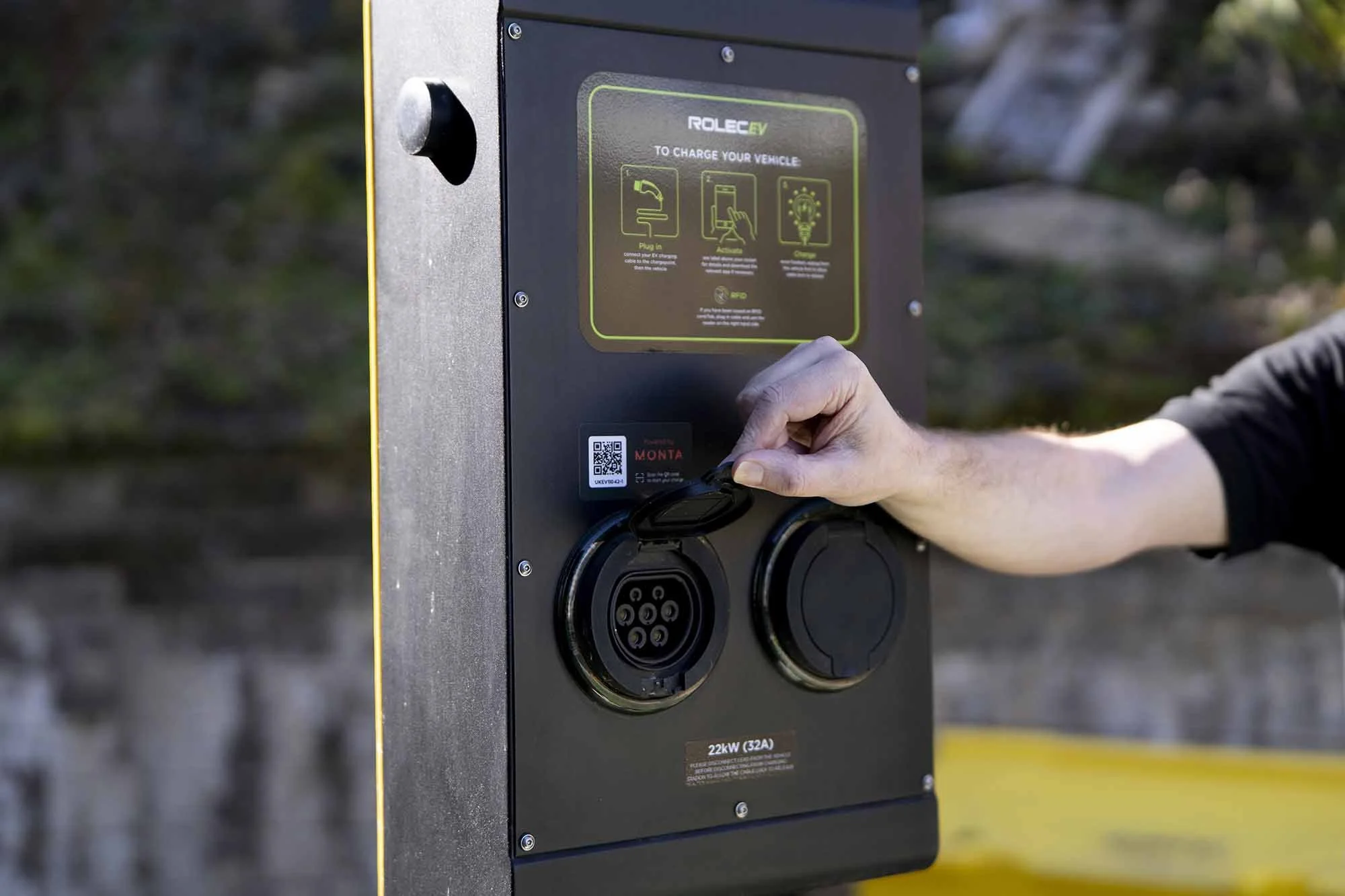 Person plugging a charging cable into an electric vehicle charging station outdoors.