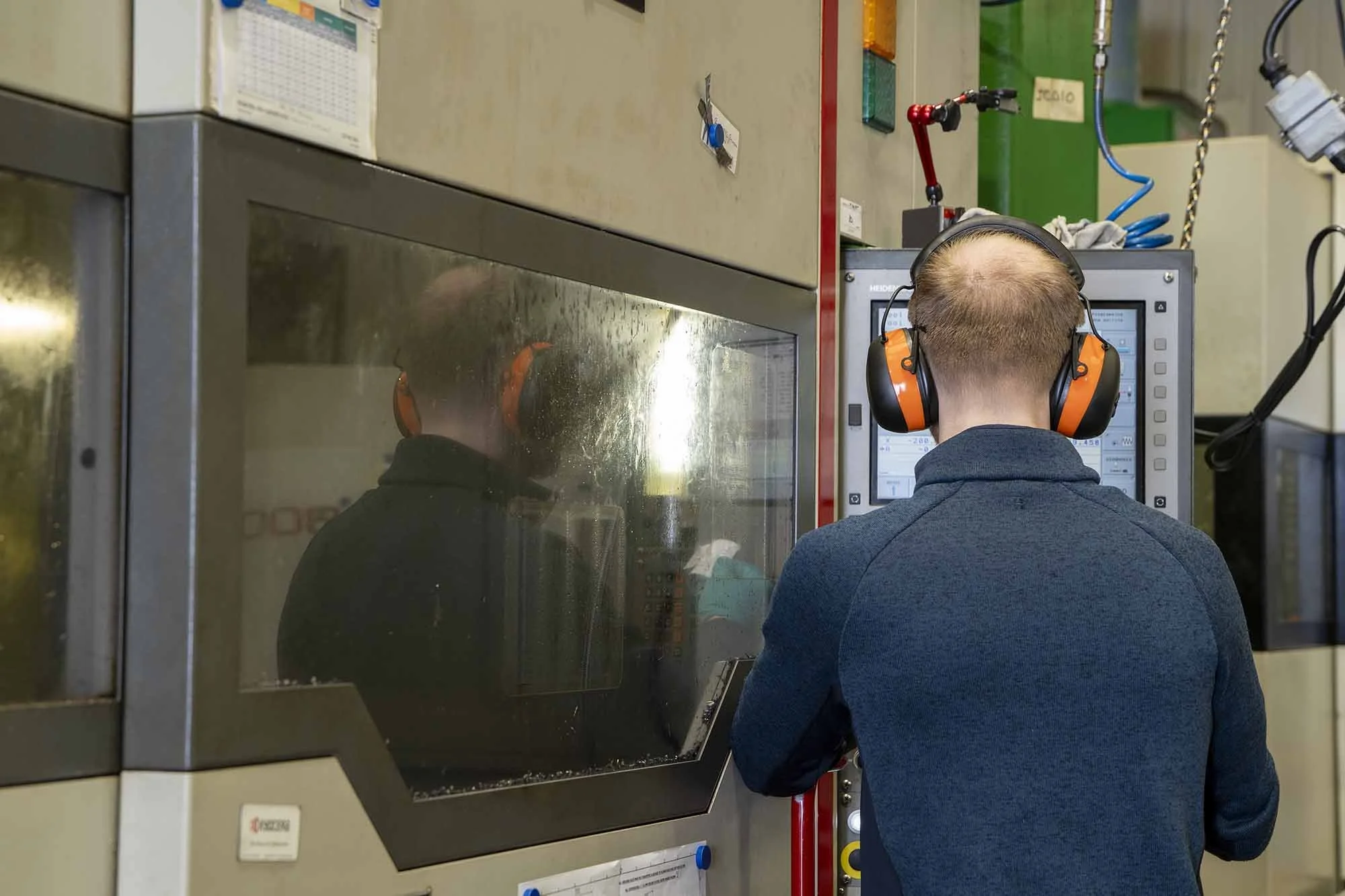 A person wearing orange and black hearing protection and a blue jacket is working on a control panel in an industrial setting. The person is looking at a digital display with various gauges and controls, and their reflection can be seen on the glass window of the machinery.