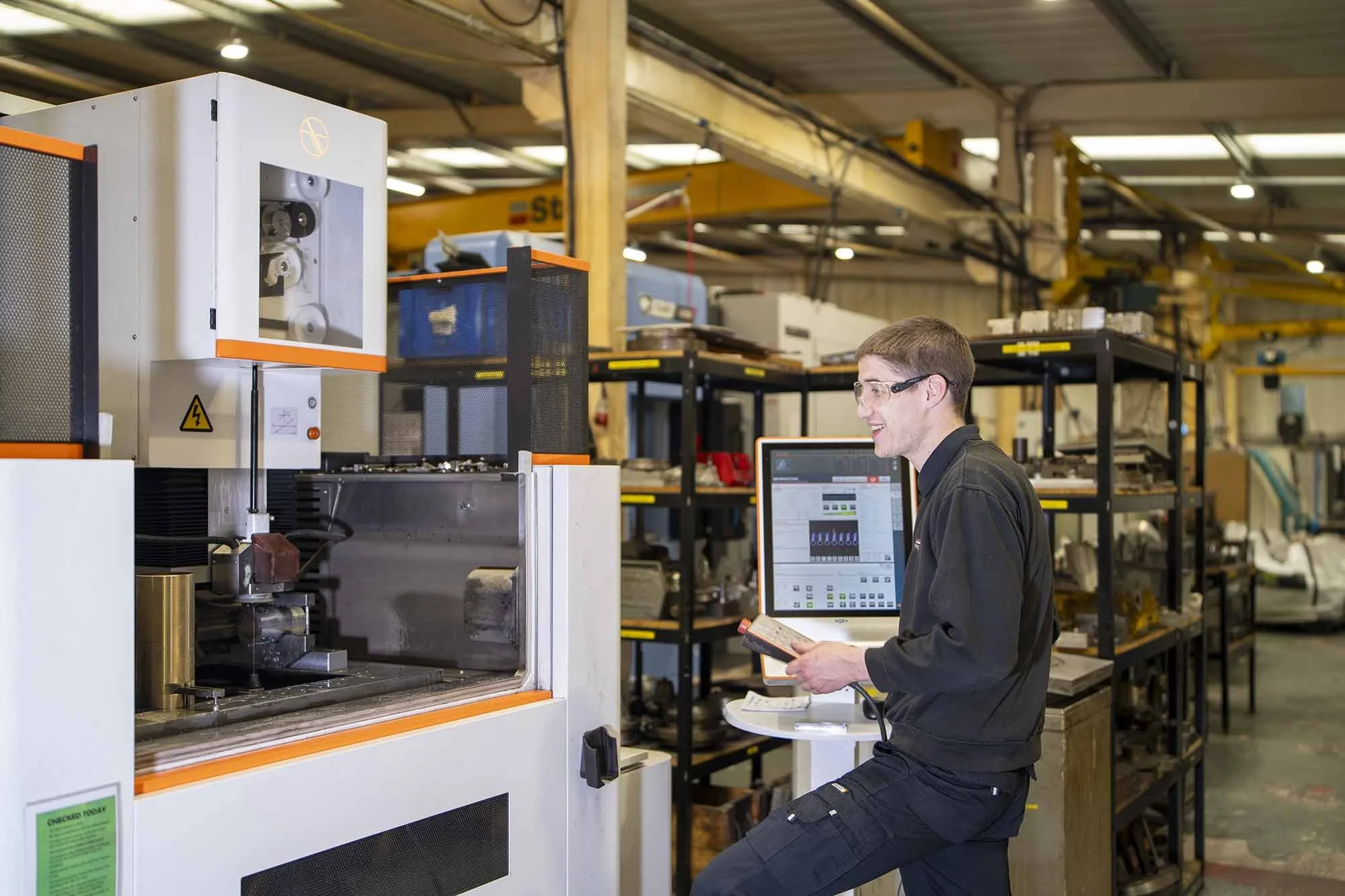A man in safety glasses operating a large industrial machine in a warehouse or manufacturing setting, with shelves of tools and equipment in the background.