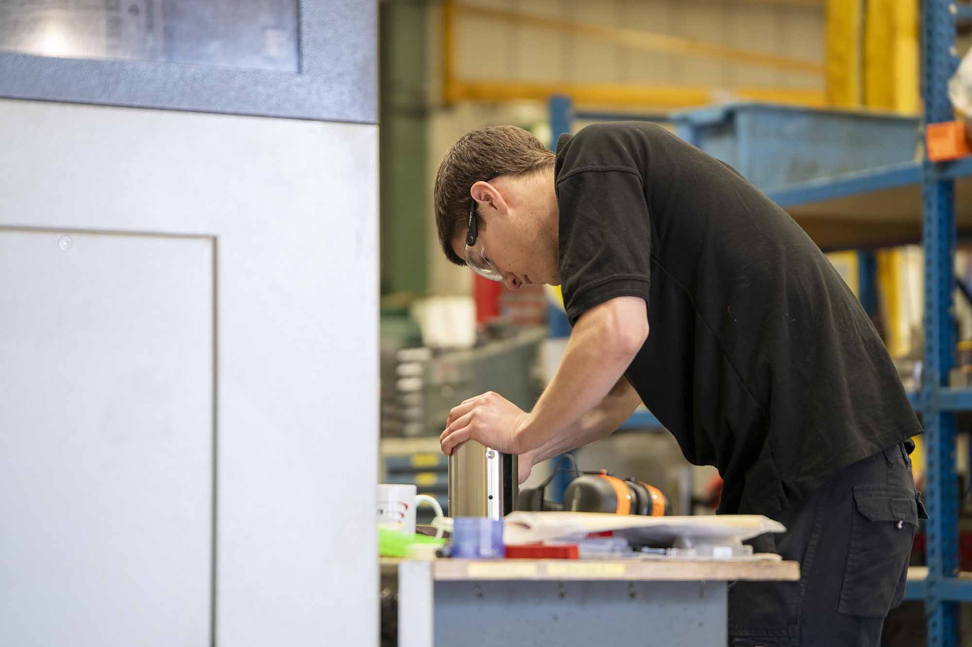 A man wearing safety glasses working with tools at a workbench in a workshop or factory setting.