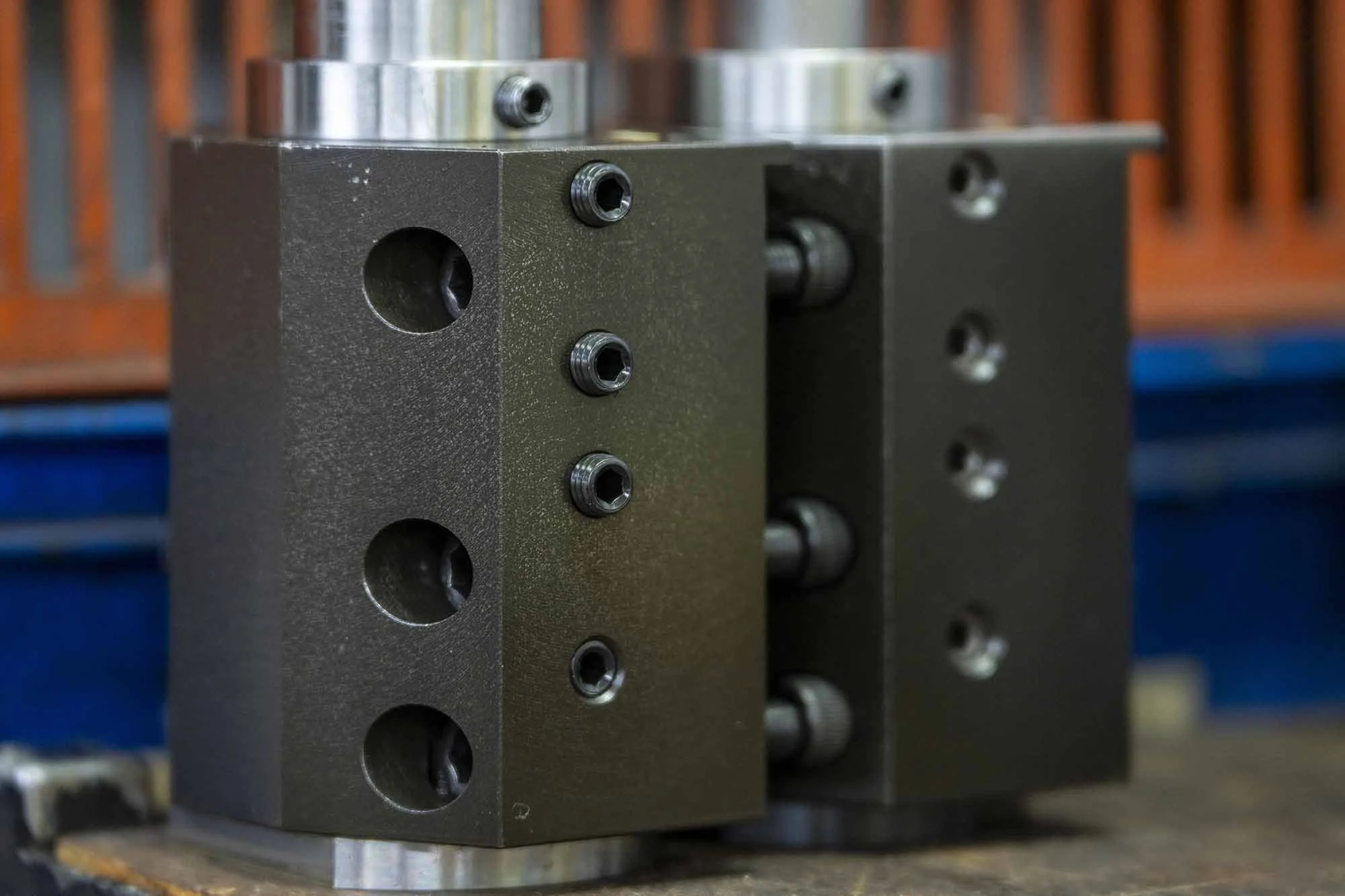 Close-up of a black industrial hydraulic valve block with multiple ports and threaded holes, mounted on a workbench with a wooden background.