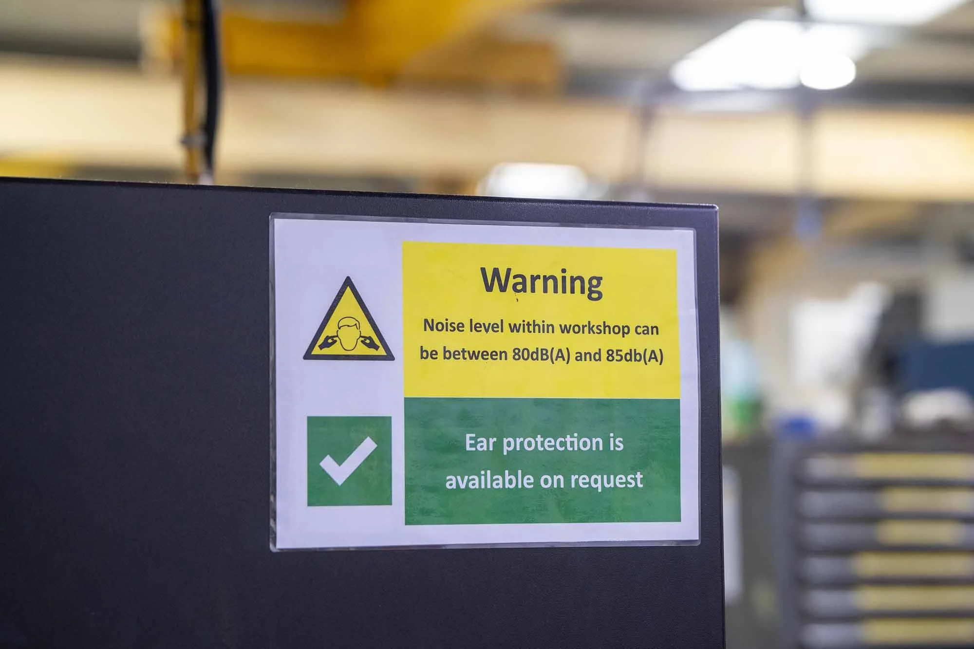 Warning sign with a yellow background and black text, indicating noise levels in a workshop between 80dB and 85dB. Green section below states ear protection is available on request.