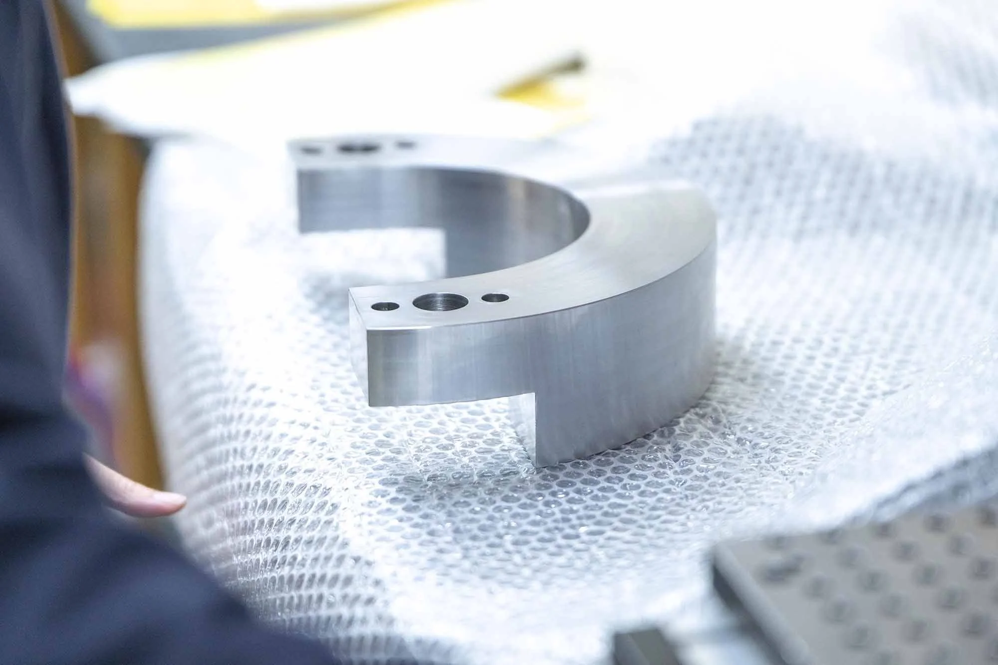 Close-up of a large, shiny metal part with multiple holes, resting on bubble wrap, likely in a manufacturing or machining setting.