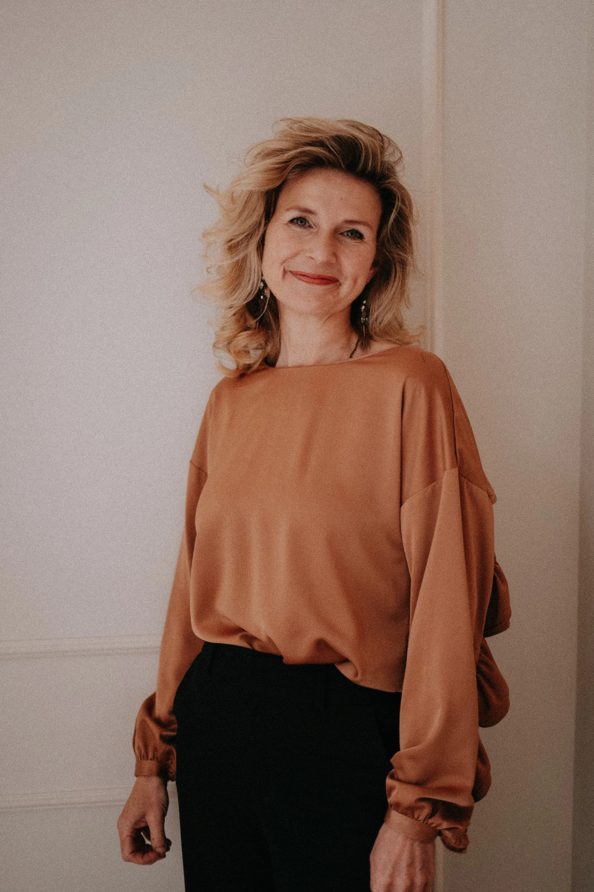 A woman with blonde, wavy hair smiling while wearing a brown satin blouse with long sleeves and black pants, standing indoors against a plain background.