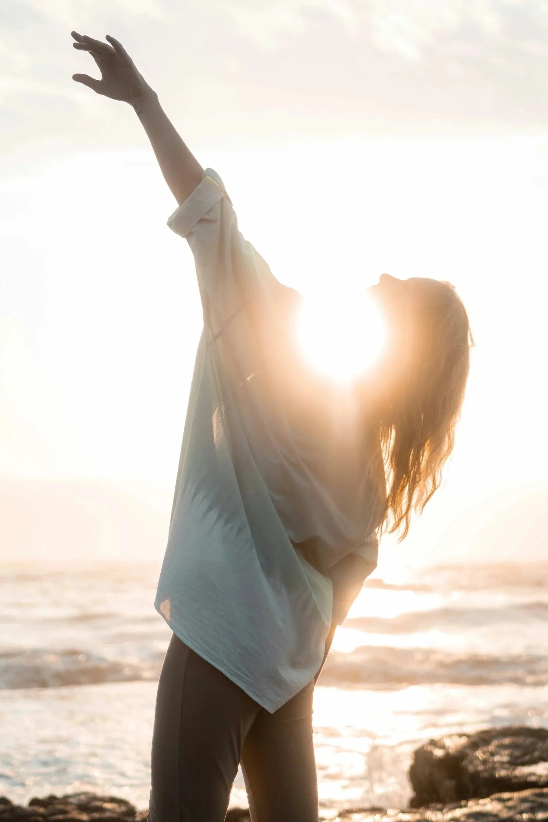 Femme debout sur la plage au coucher du soleil, levant un bras vers le ciel, silhouette éclairée par la lumière du soleil