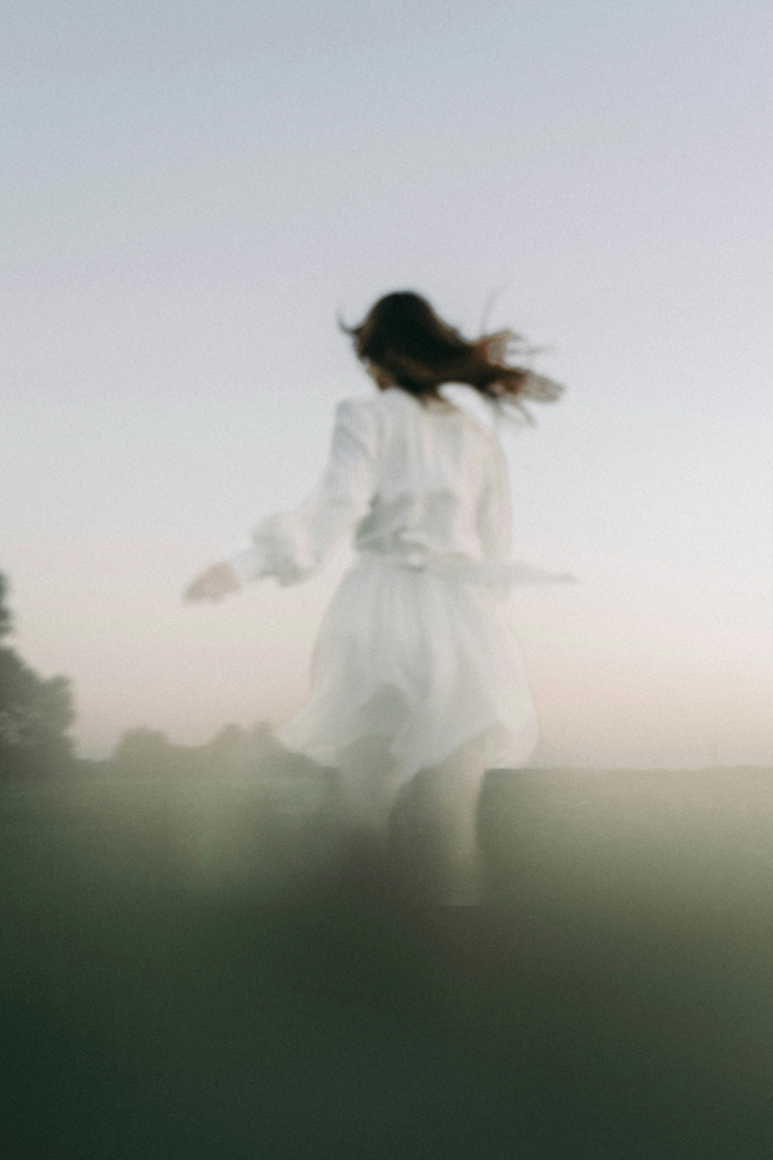 Une femme en robe blanche marche dans un paysage au coucher du soleil, avec un ciel pastel et une silhouette de forêt au fond.