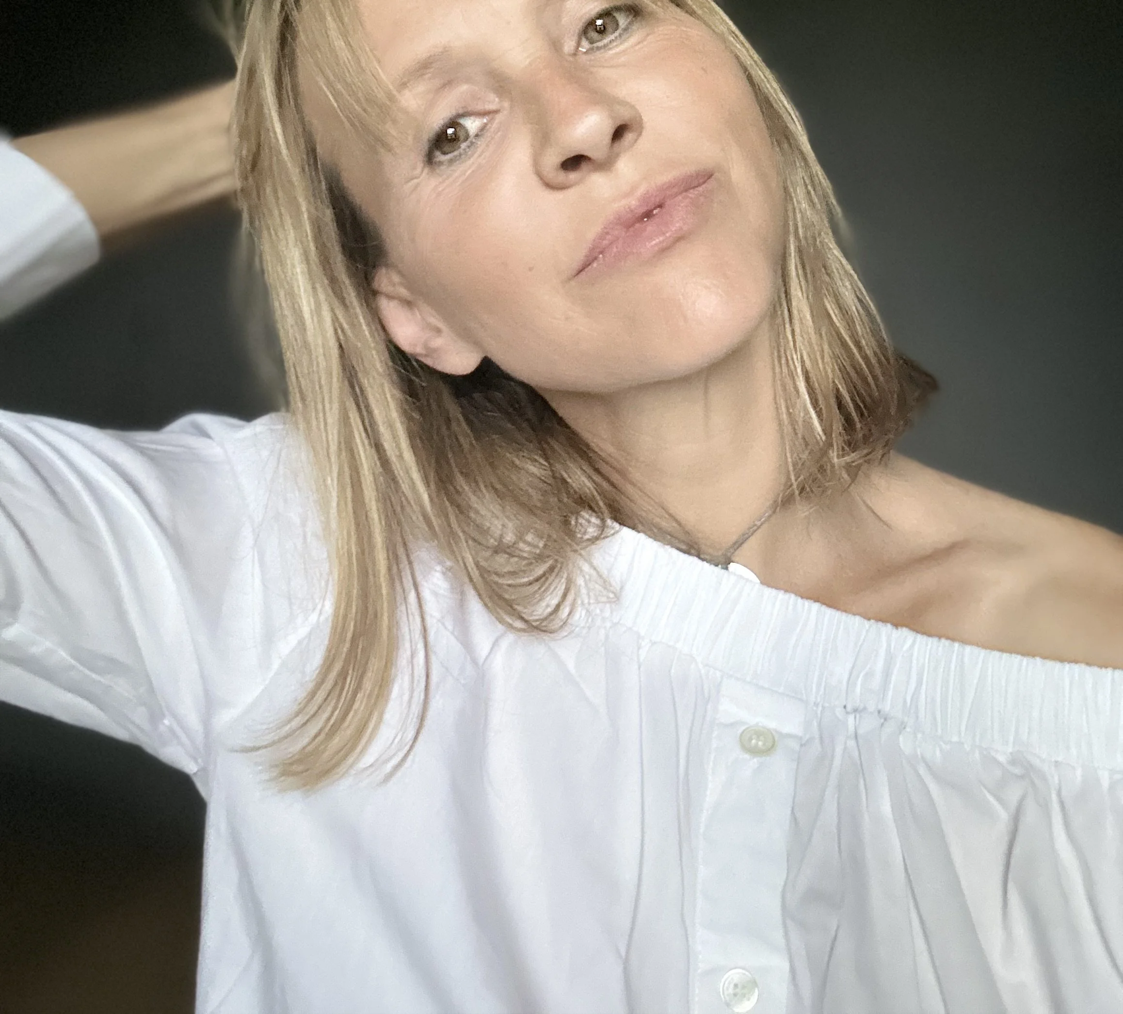 Une femme aux cheveux blonds, portant une chemise blanche, regarde l'appareil photo d'un air détendu, dans un contexte intérieur.