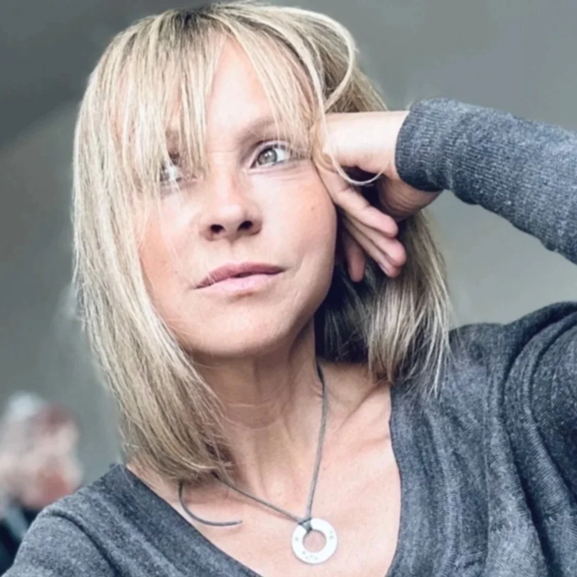 Une femme à la blondeur moyenne, les cheveux légèrement ondulés, posant pour un selfie. Elle a un regard tranquille et porte un vêtement gris ainsi qu'un collier avec un pendentif circulaire.