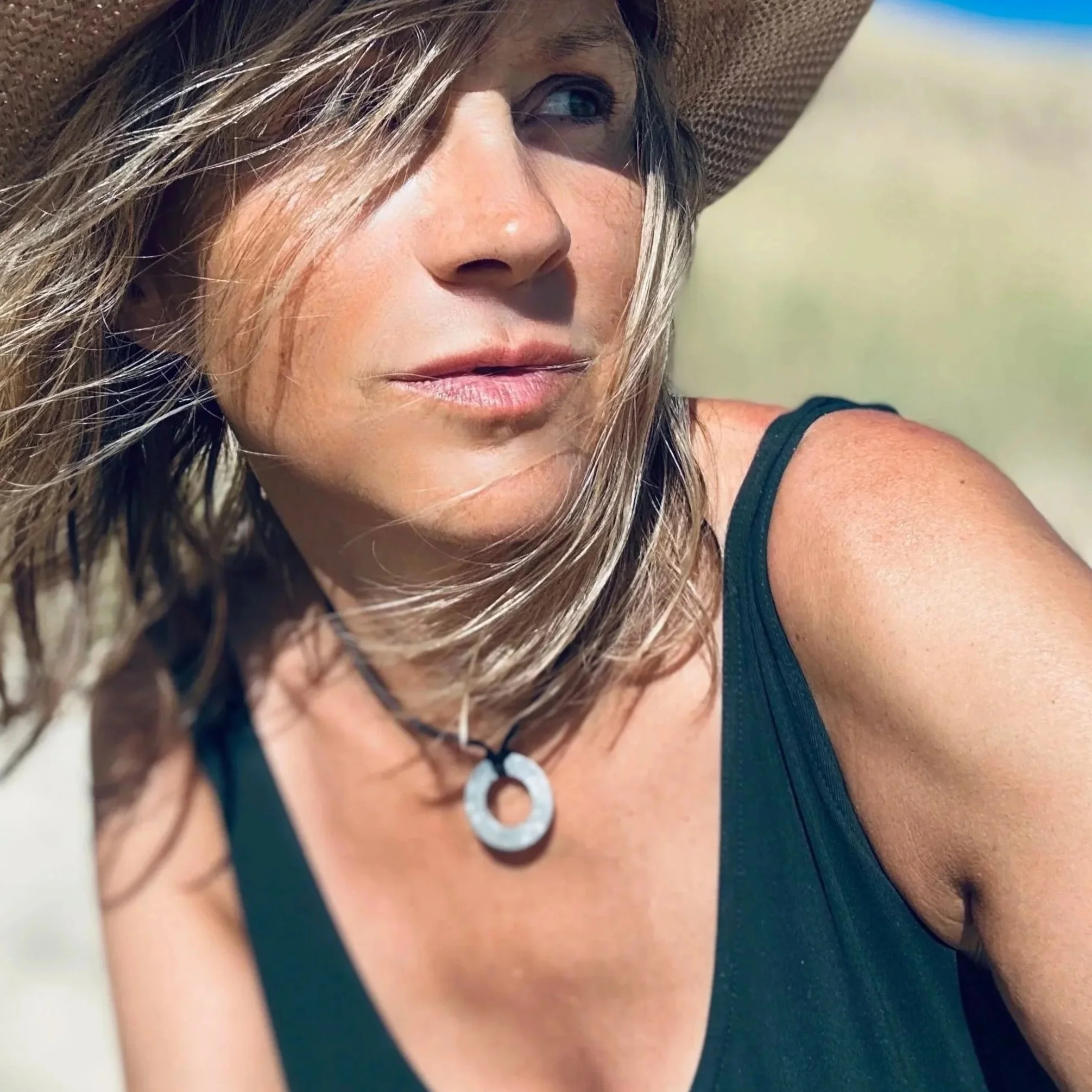 Femme avec chapeau, cheveux bruns, porte un colier avec un pendentif, portant un haut noir, à l'extérieur par une journée ensoleillée.