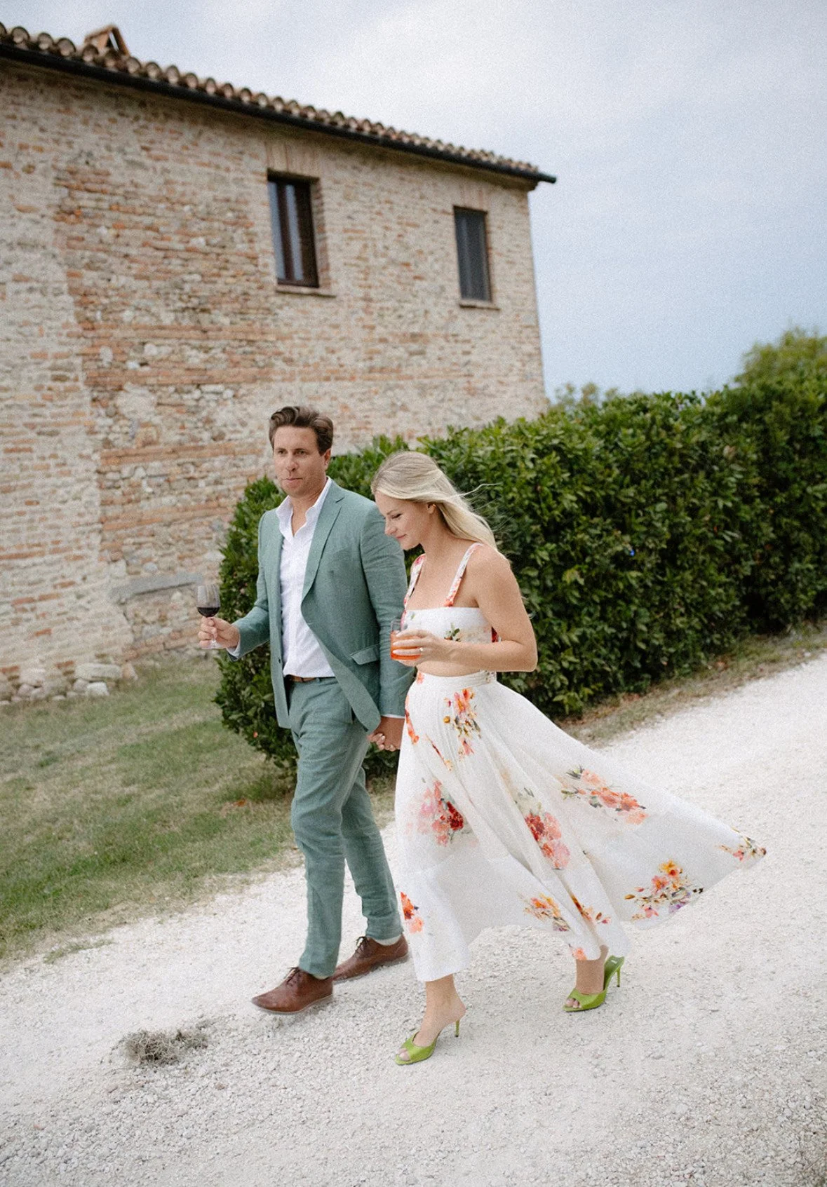 A couple walking outdoors on a gravel path, holding hands and drink. The woman is wearing a floral matching set and green heels, the man is in a light suit and brown shoes. They are at their welcome party in Italy, a destination wedding.