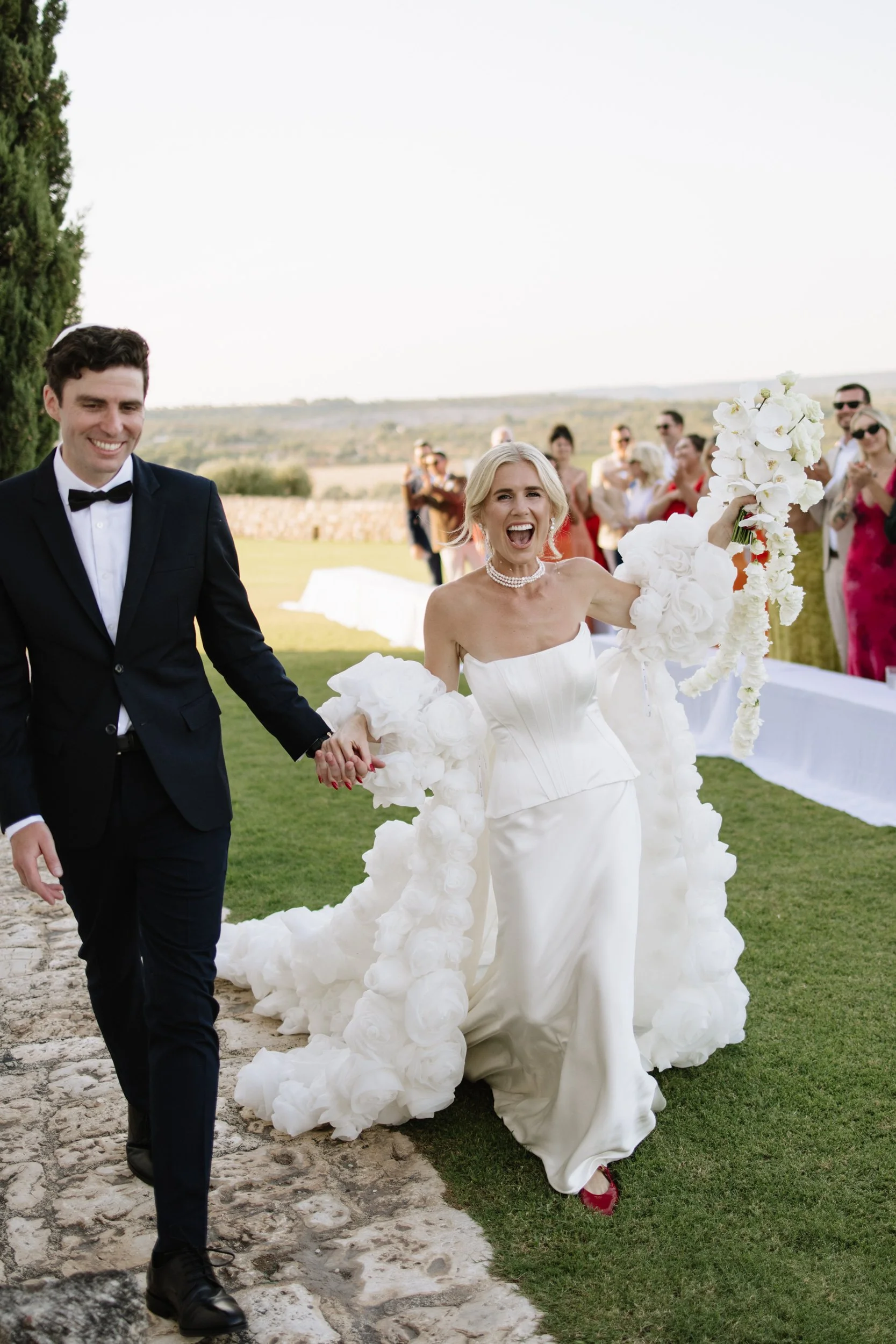 Bride and groom walking down the aisle at Italian summer wedding, wearing statement floral cape and corset gown