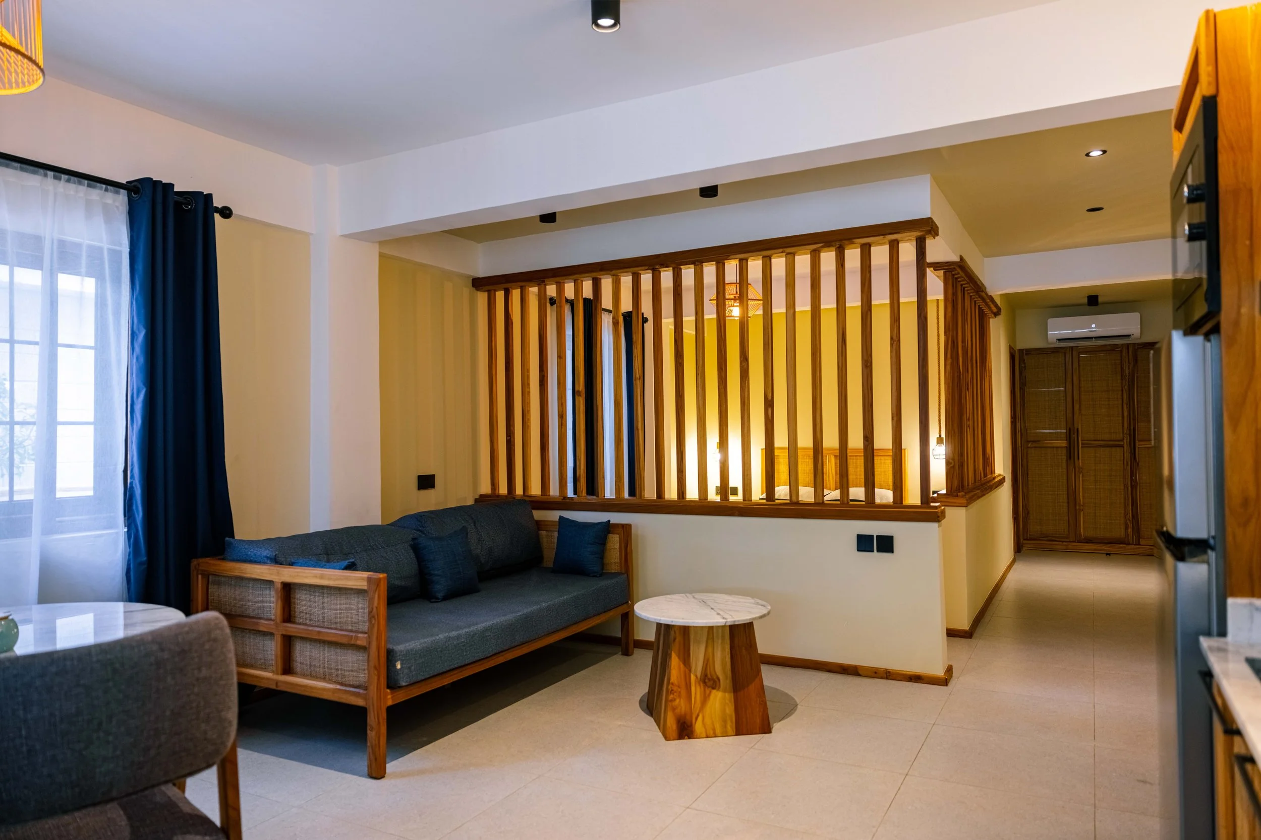 Living room with a gray sofa, a wooden coffee table, and large windows with blue curtains. A wooden partition separates the living space from the bedroom area, which has a bed and a wall-mounted air conditioning unit.