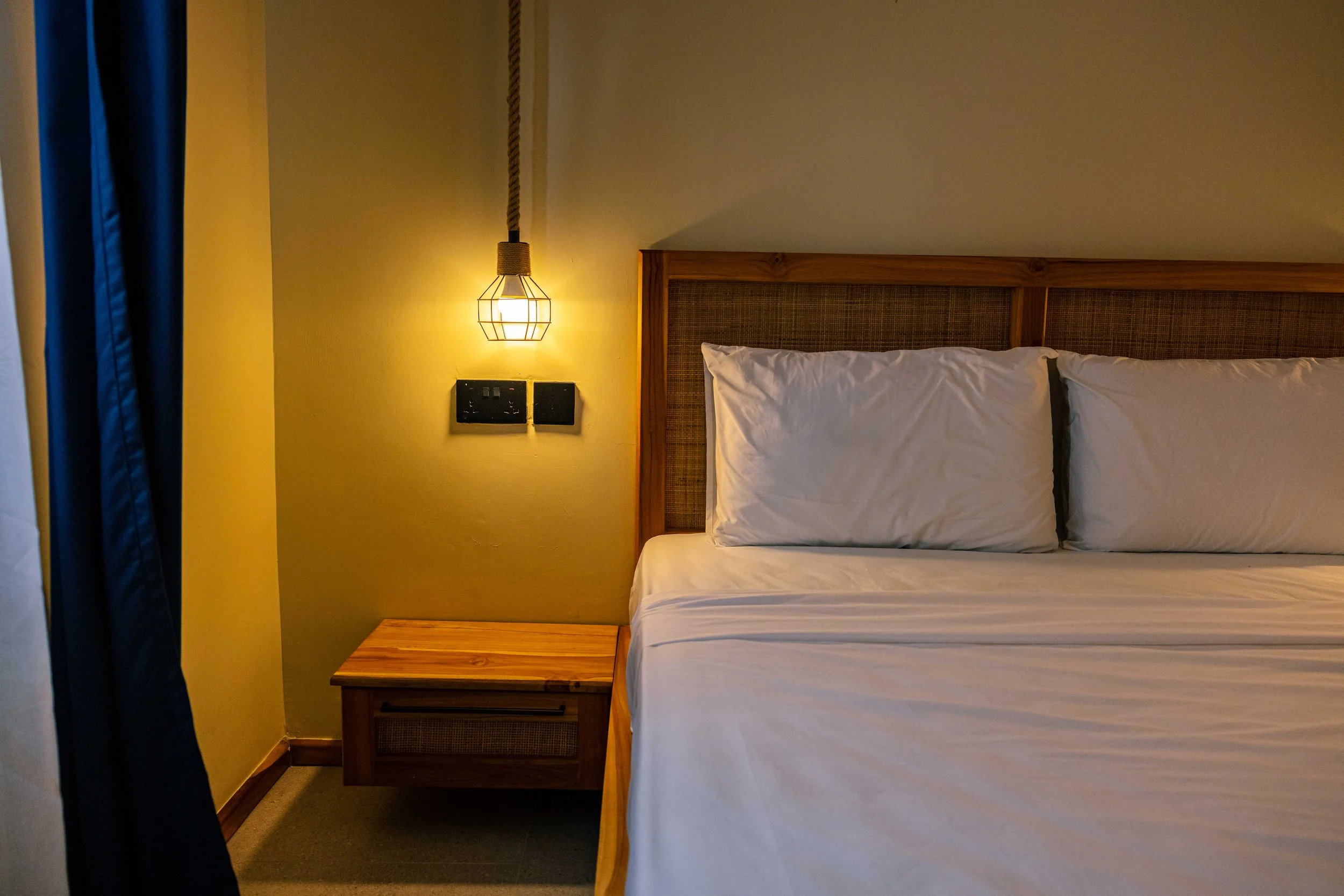 A bedroom with a bed featuring white sheets and pillows, a wooden headboard, a bedside table, and a geometric pendant light fixture on a yellow wall.