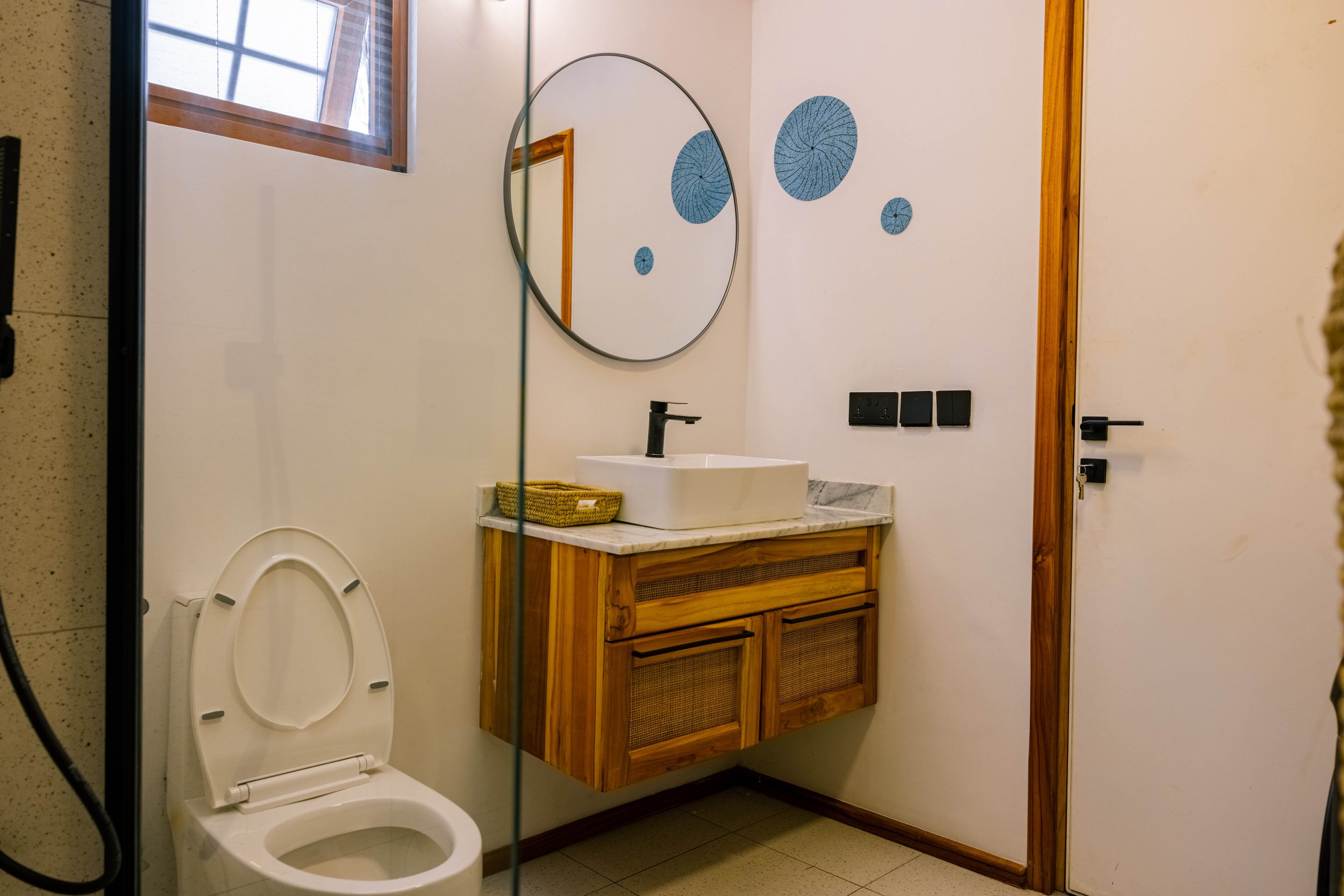 A bathroom with a white toilet, a wooden vanity with a white sink and black faucet, a large round mirror, decorative blue wall art, and a window with wooden trim.