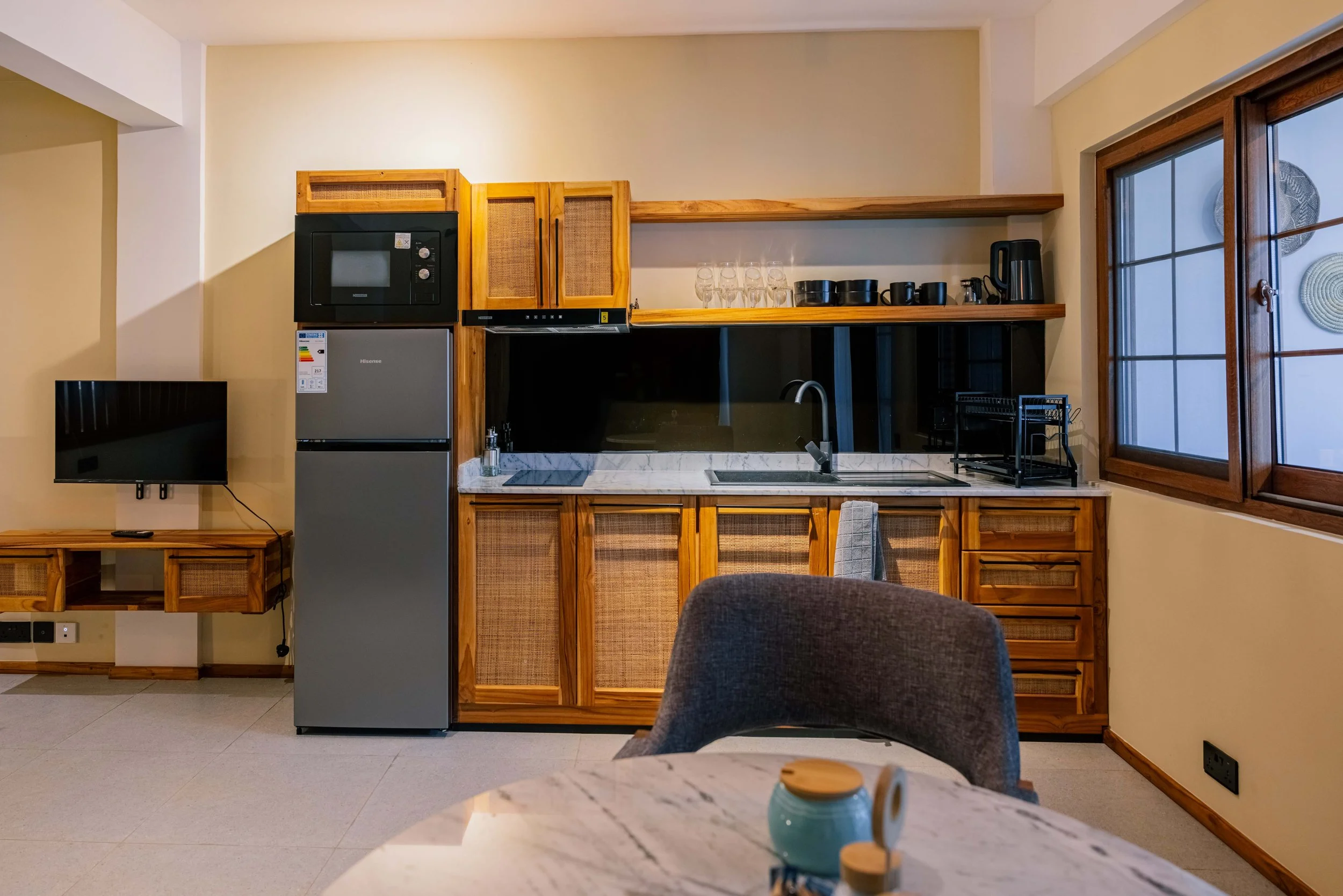 A kitchen with wooden cabinets, a marble countertop, a microwave, a mini refrigerator, shelves with glasses and cups, a window, and a chair.