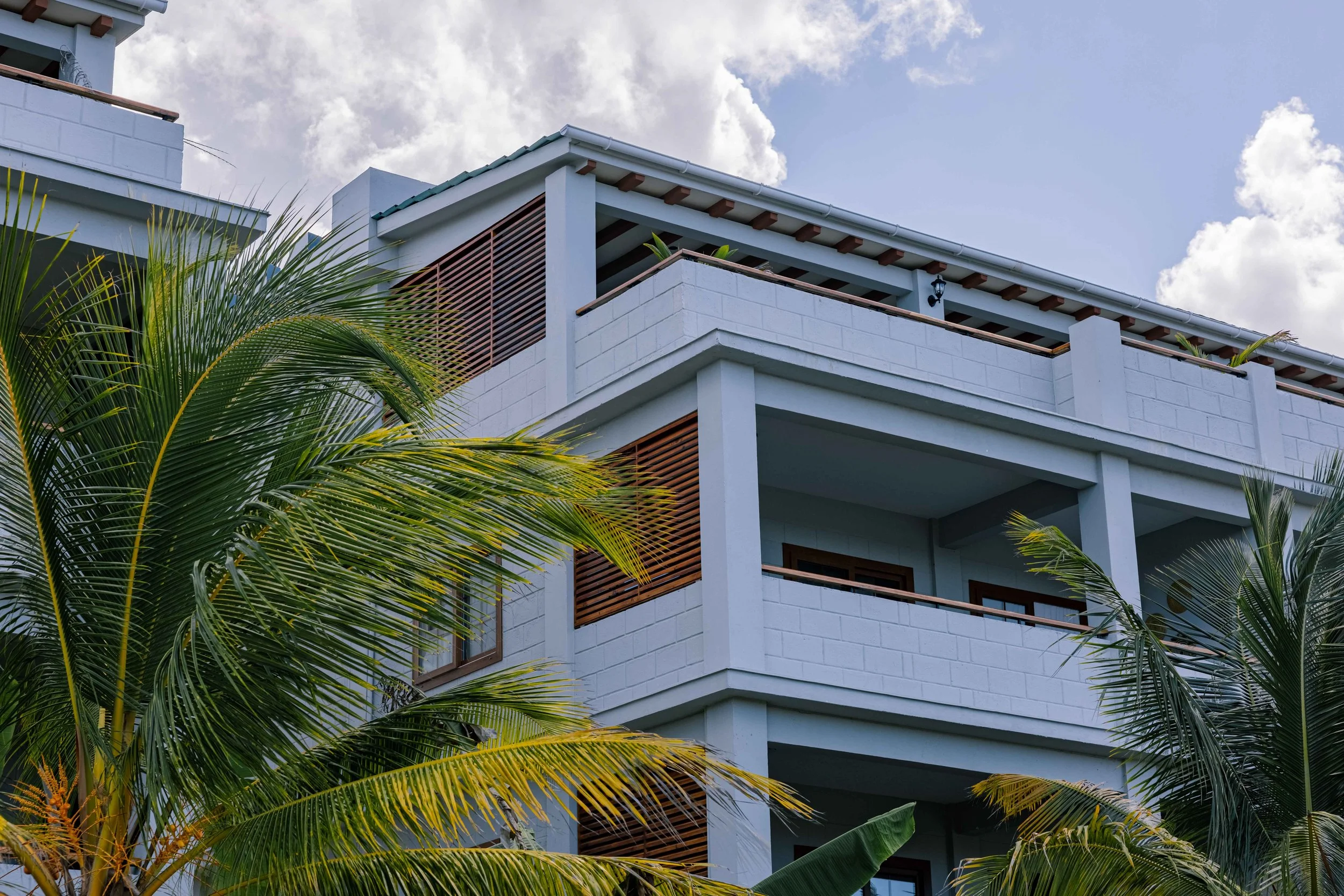 A modern multi-story building with white walls and wooden accents, surrounded by lush green palm trees, under a partly cloudy sky.