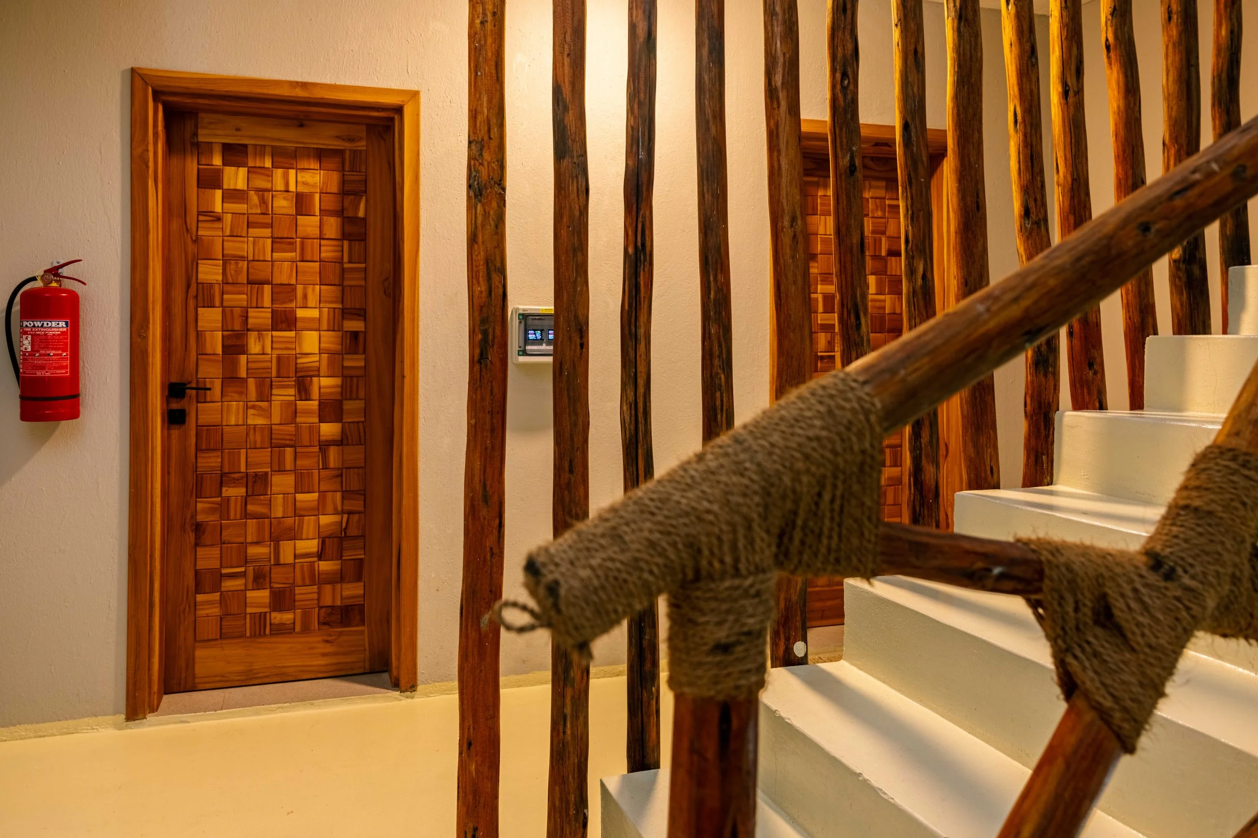 Interior of a house showing a staircase with a woven rope railing, two wooden doors with a geometrical pattern, a fire extinguisher on the wall, and a small wall-mounted control panel.