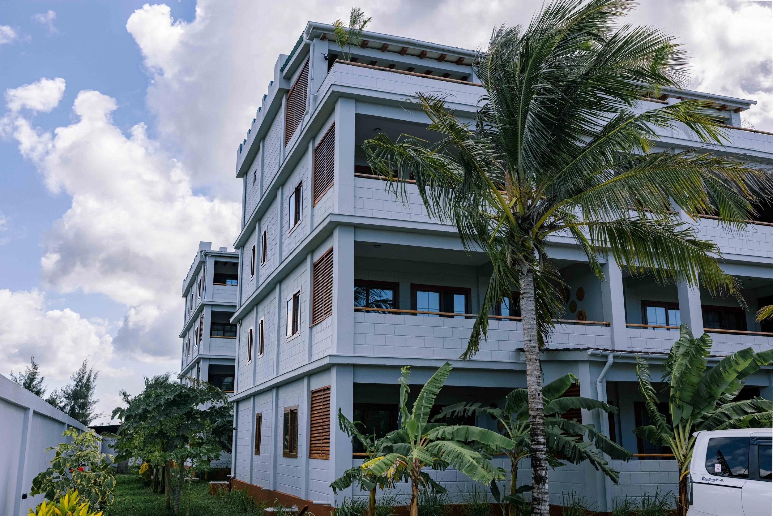 A modern multi-story building with balconies, surrounded by lush green tropical plants and palm trees, under a partly cloudy sky.