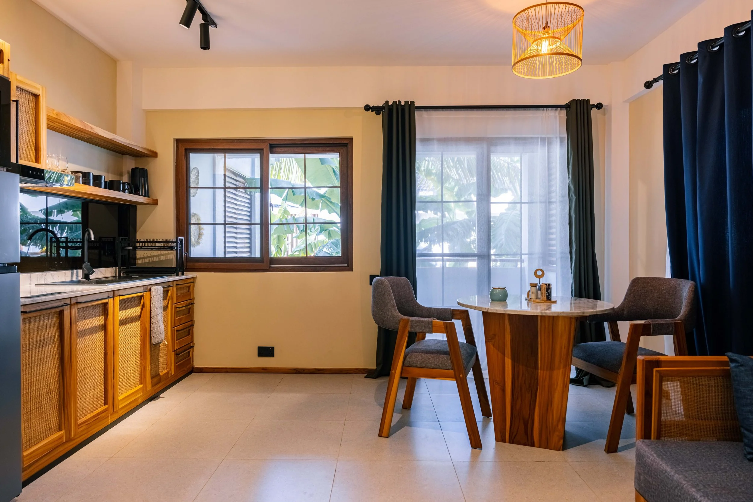 A cozy dining area with a round marble table surrounded by four upholstered chairs, large windows with sheer curtains and dark drapes, a ceiling light fixture, and a kitchen in the background with wooden cabinets, open shelves, and a small window wit