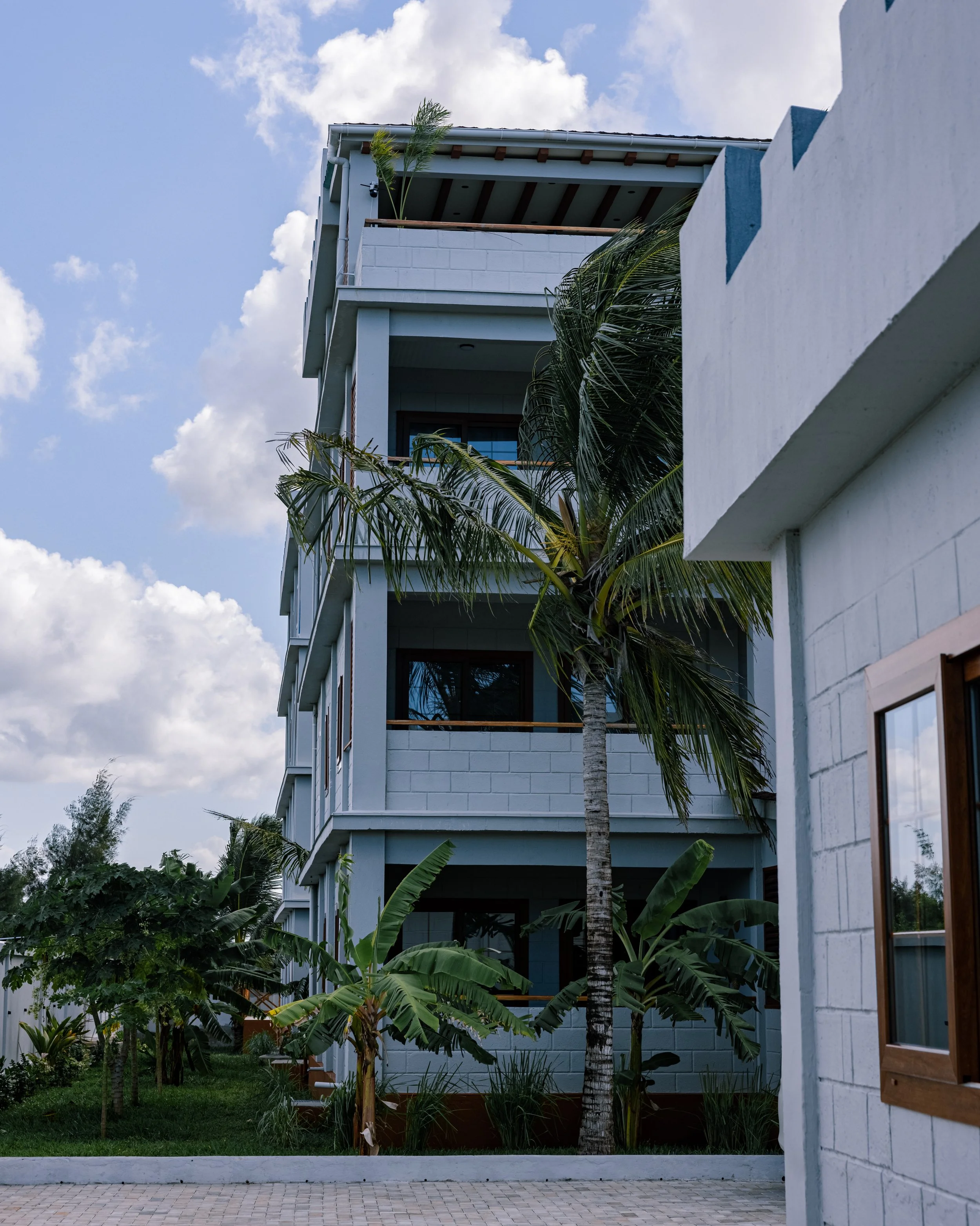 Multi-story modern building with balconies, surrounded by palm trees and lush greenery, under a partly cloudy sky.