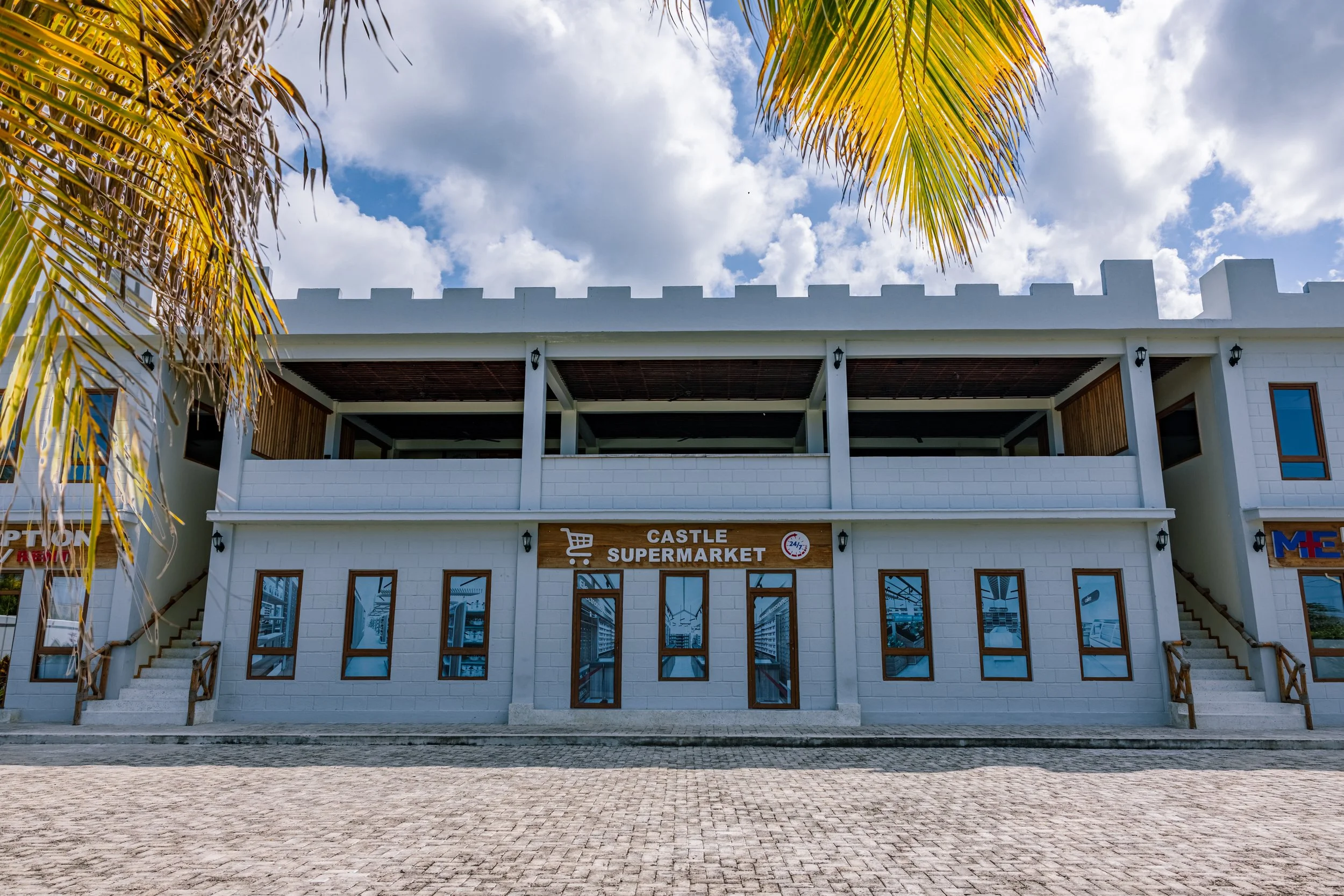 Front view of a white building with a sign reading 'Castle Supermarket' and a shopping cart icon, under a partly cloudy sky with palm tree leaves at the top.