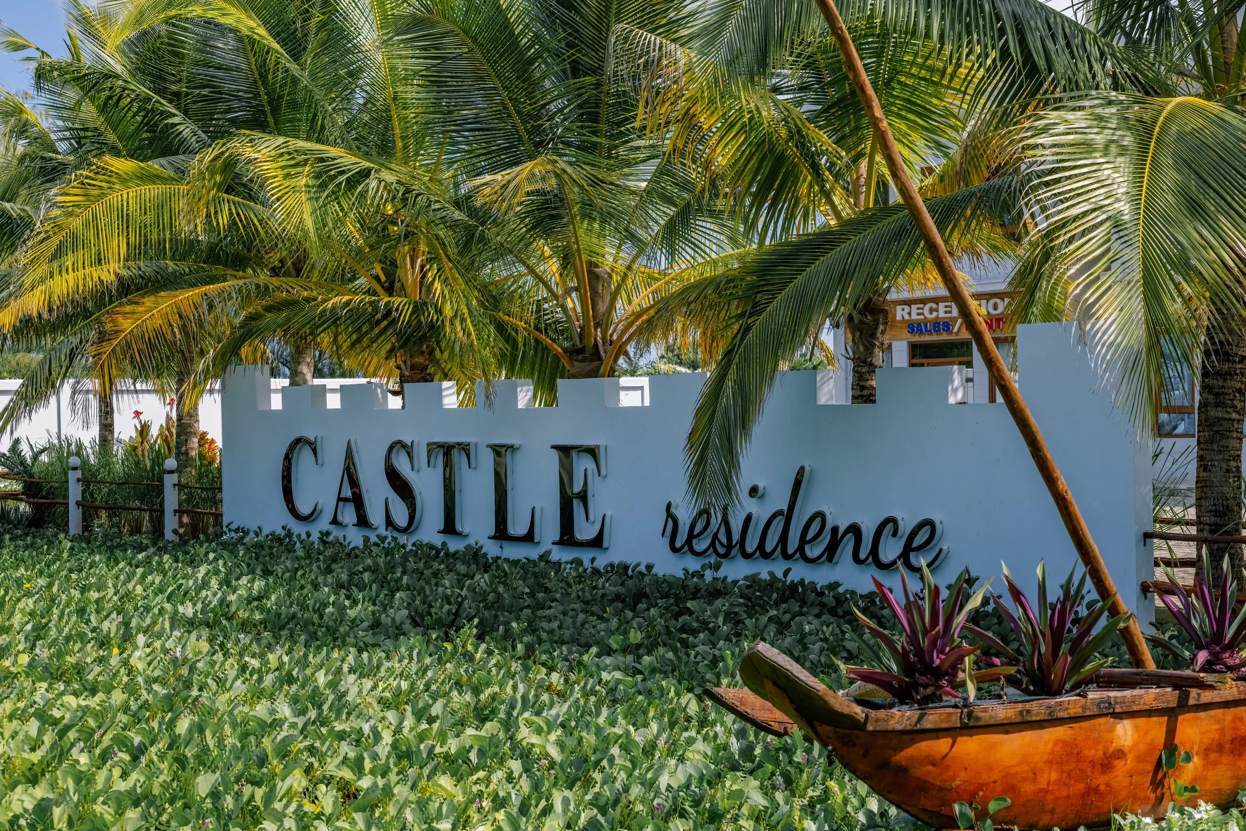 A white sign reading 'Castile Residence' surrounded by lush green plants, palm trees, and a blue sky, with a wooden boat filled with vibrant flowers in the foreground.