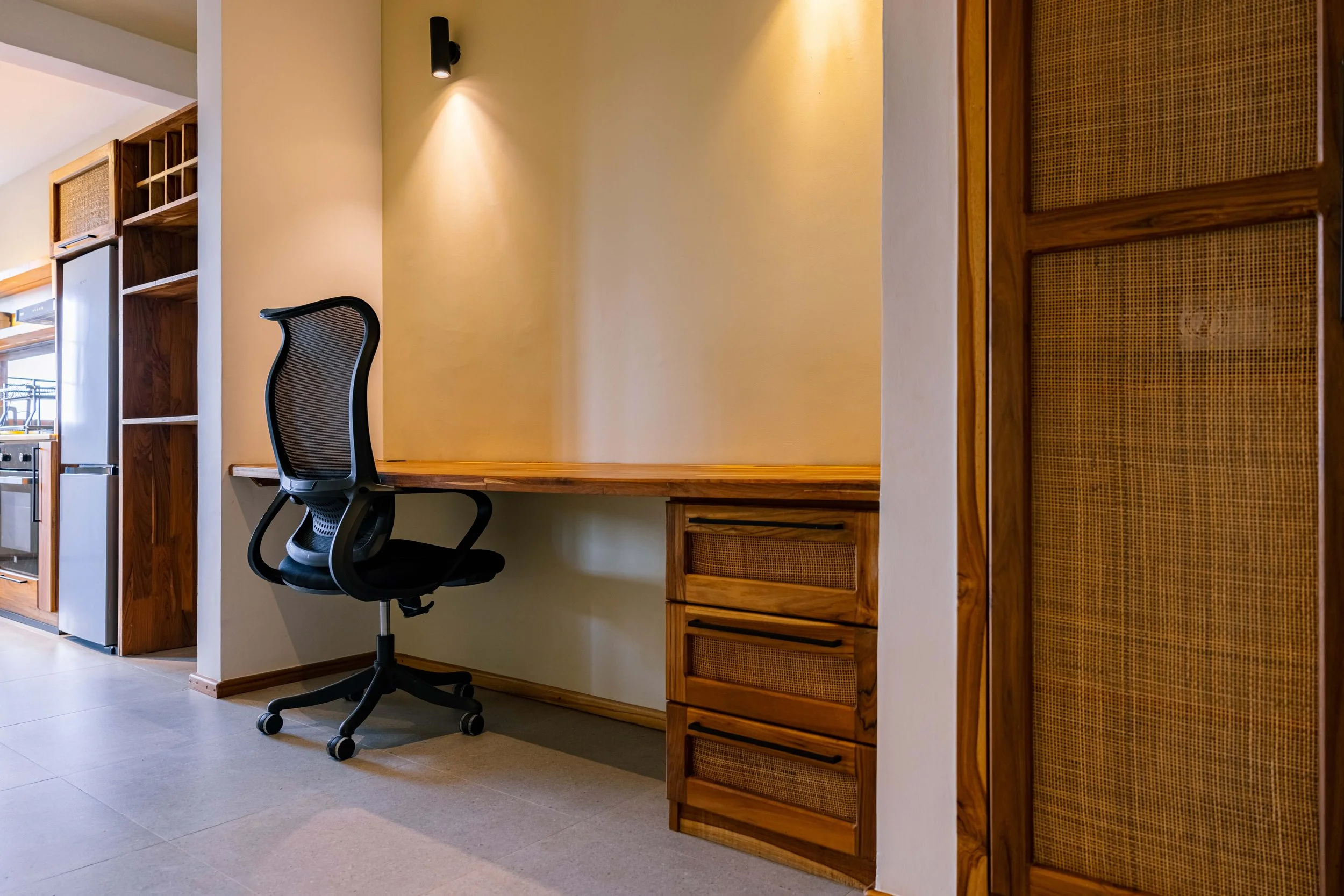 Home office with a wooden desk, black ergonomic chair, and wooden shelving on a beige wall.