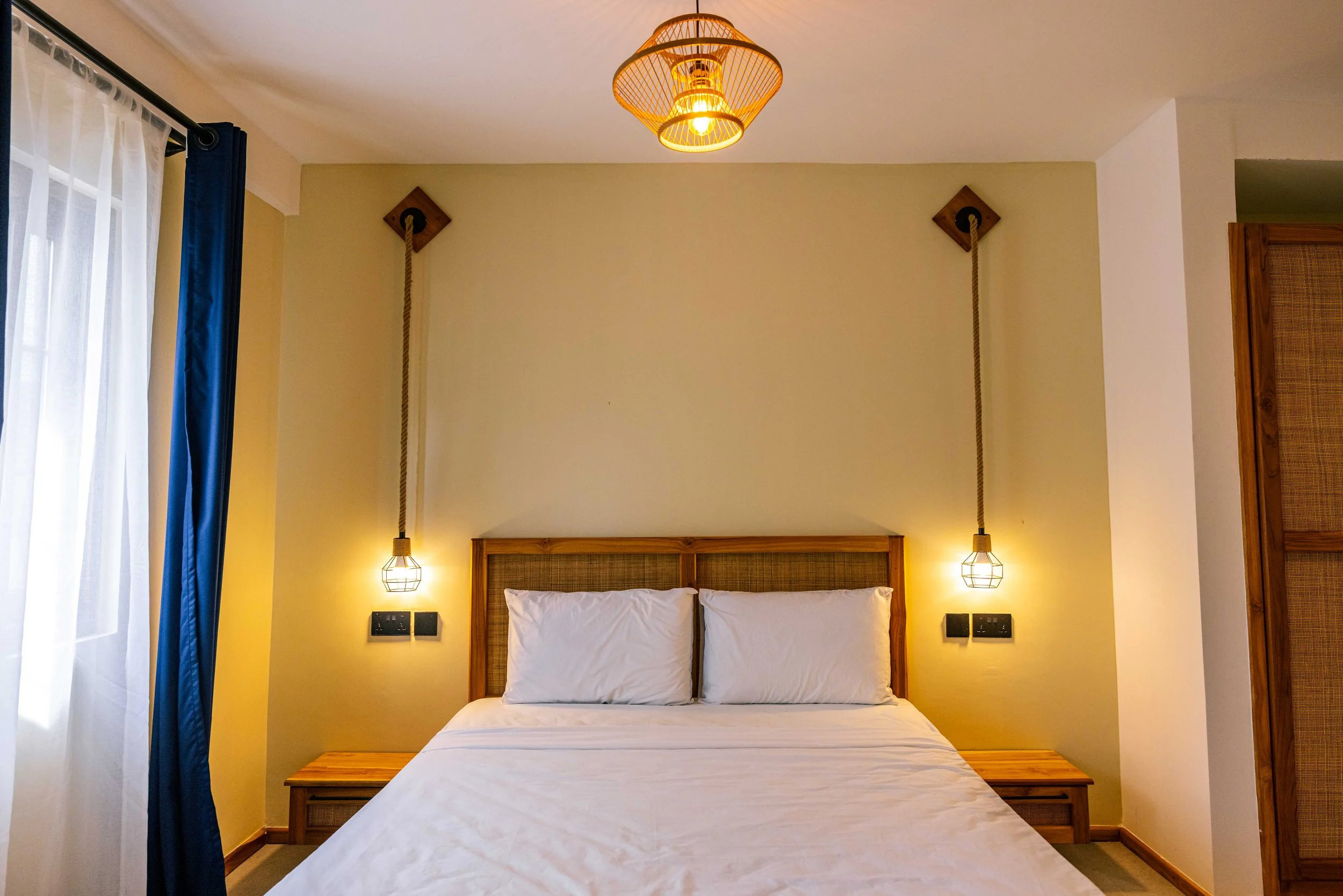 A bedroom with a double bed, white pillows, and a wooden headboard. Two hanging lamps on either side of the bed, a window with white curtains and blue drapes on the left, and a yellow wall with two ropes attached to wooden mounts.