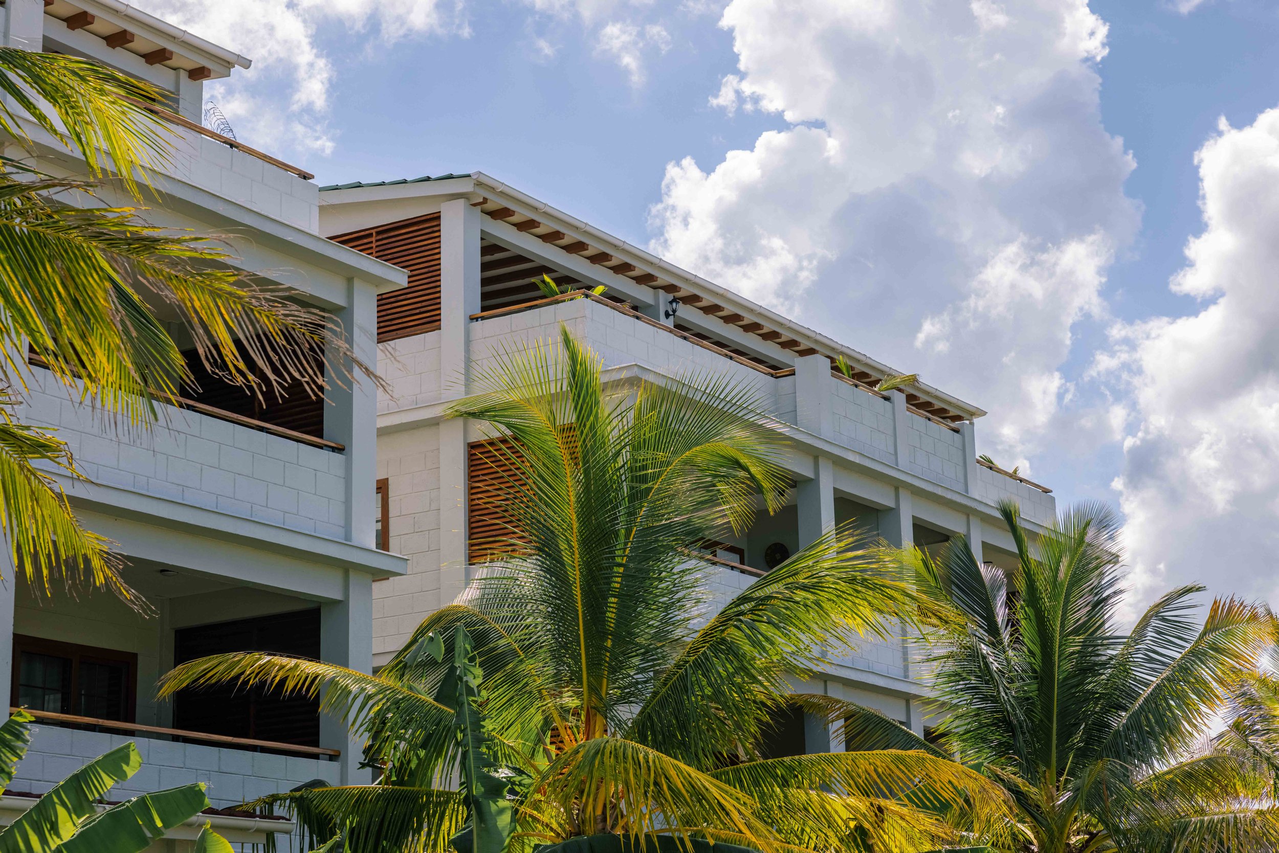 Modern white multi-story building with balconies and wooden accents, surrounded by green palm trees, under a partly cloudy sky.