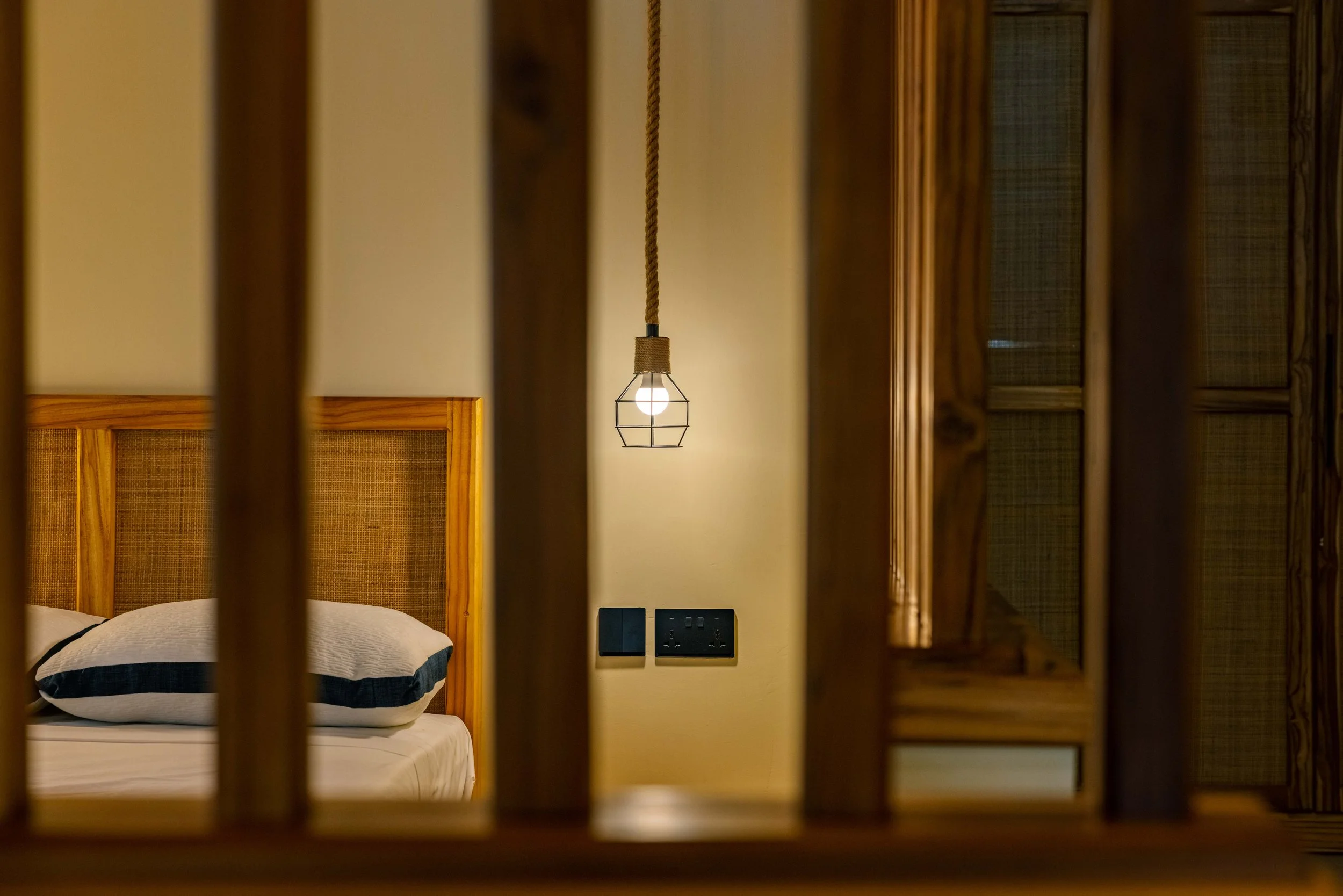 View through wooden slats of a bedroom with a bed, pillows, a headboard, a glowing pendant light, and electrical outlets on the wall.
