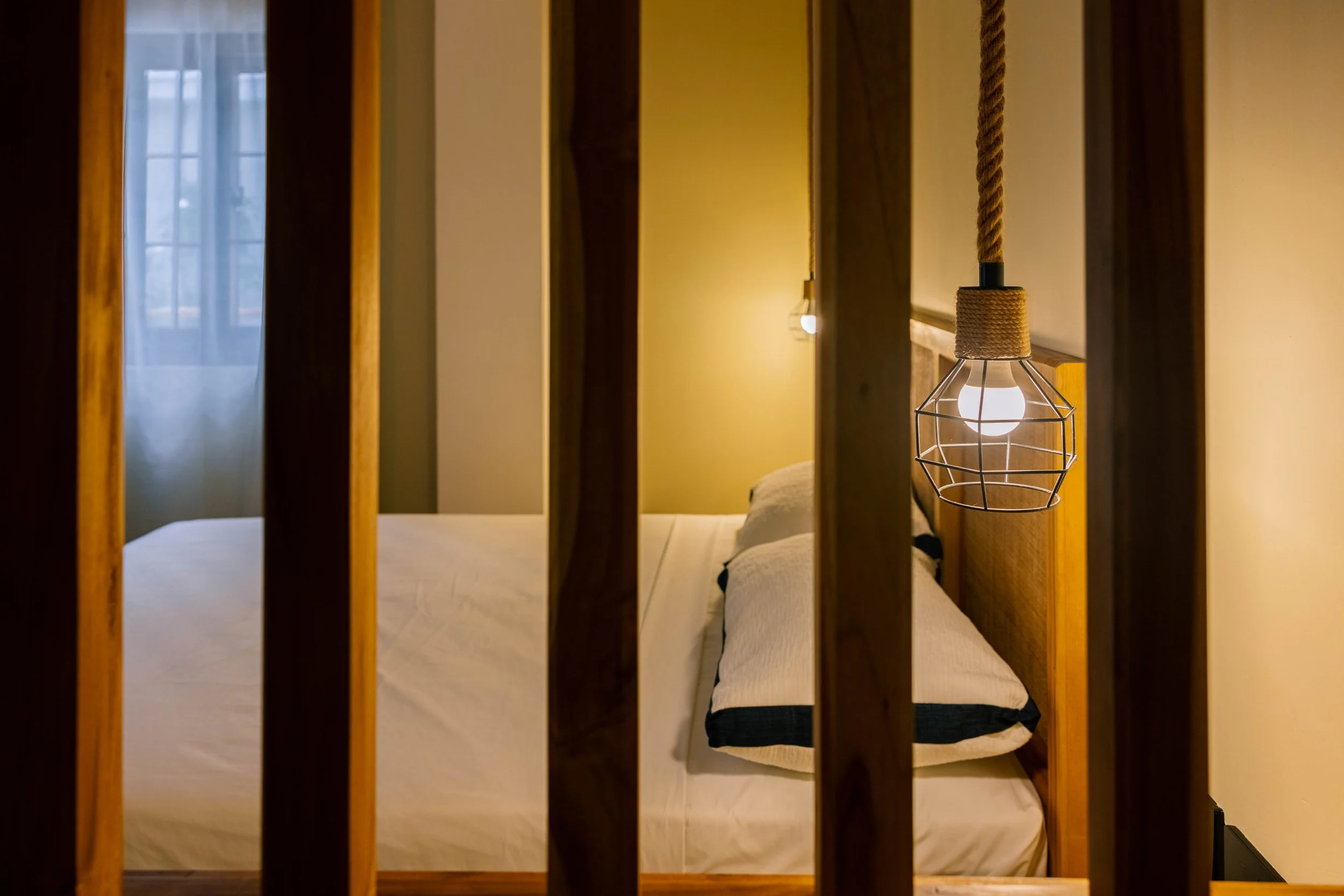 A bedroom with a bed behind wooden slats, a hanging lamp with a wireframe shade, and a window with curtains, illuminated by soft yellow lighting.