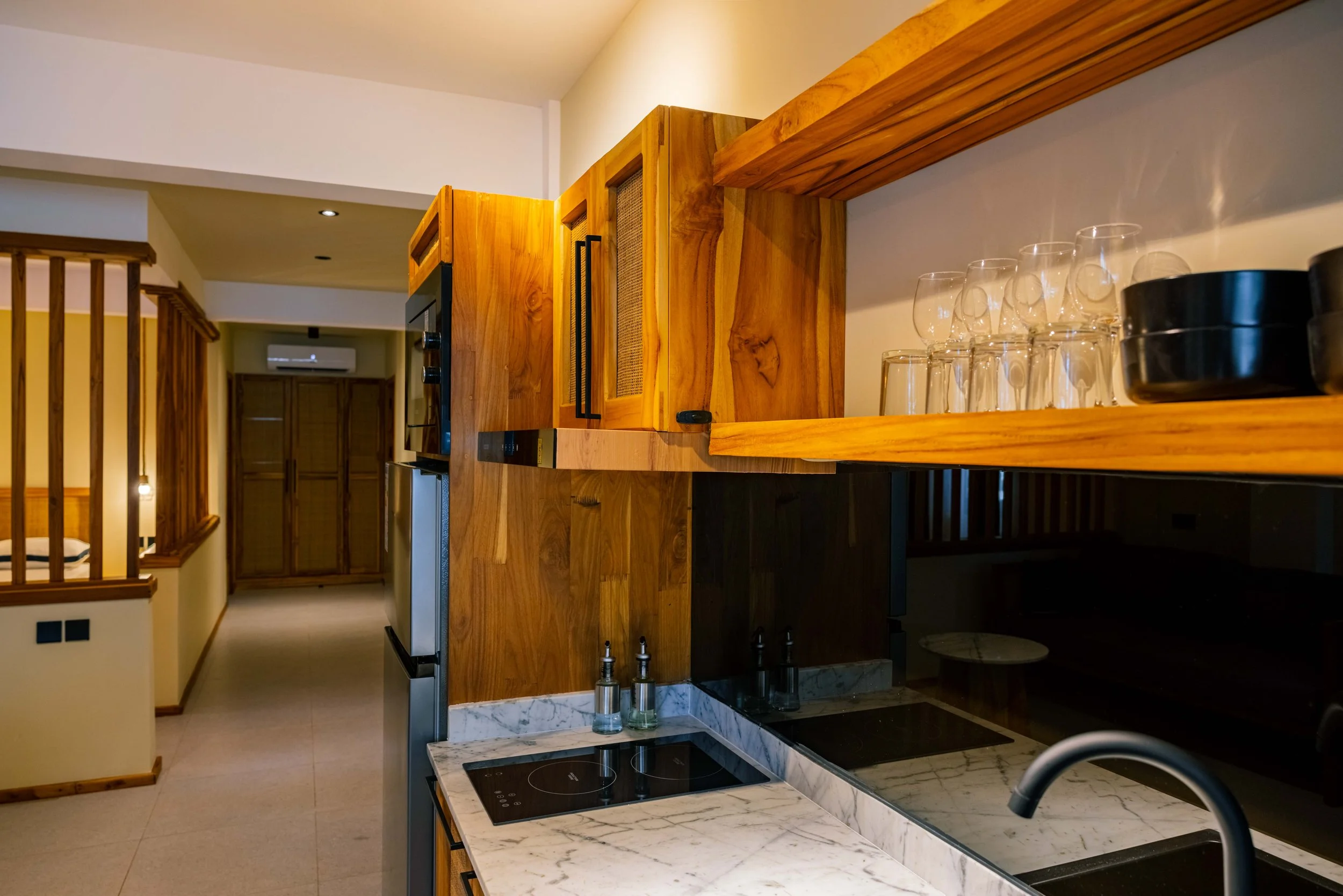 A kitchen with wooden cabinets, a marble countertop with an electric cooktop, a black sink, and a rack with glasses and bowls. Wooden railings and a hallway with an air conditioning unit in the background.