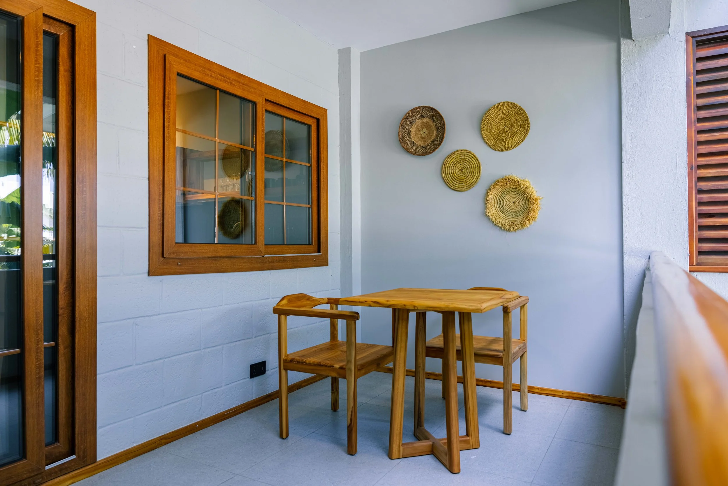 A corner of an outdoor patio or balcony with a white brick wall, wooden-framed glass windows, a small wooden table, and woven decorative wall hangings.