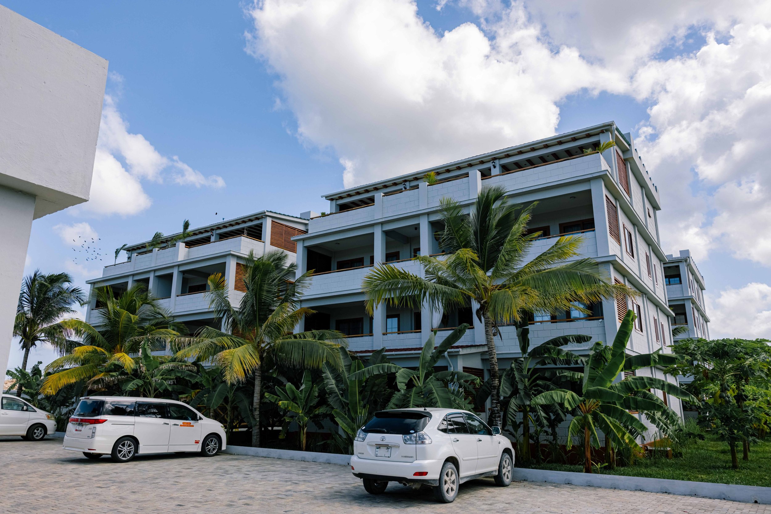 A modern white multi-story hotel building surrounded by tropical palm trees under a partly cloudy sky, with four parked cars in the foreground.
