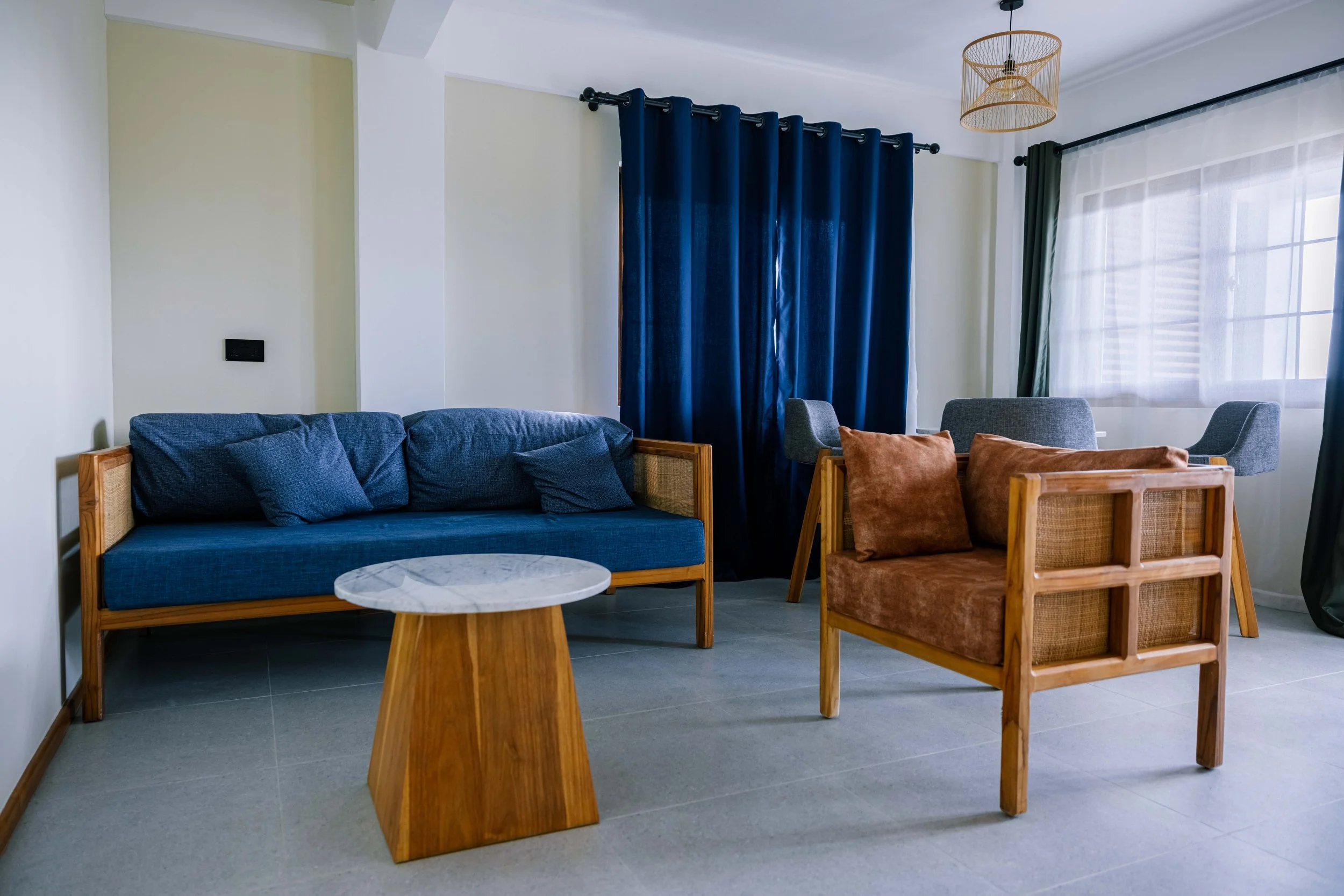 Living room with blue sofa, wooden armchair with brown cushions, small round white marble coffee table, dark blue curtains, white sheer curtains, hanging light fixture, and large window.