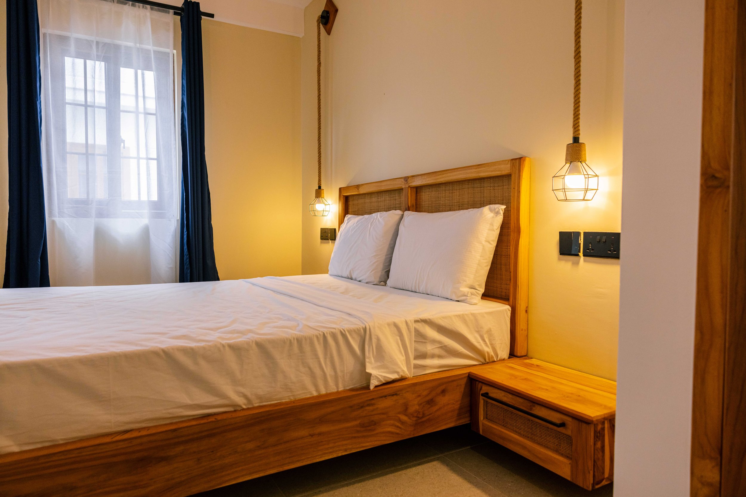 A neatly made bed with white bedding and pillows in a hotel room, featuring a wooden headboard, hanging pendant lights, a window with blue curtains, and a small wooden bedside table.