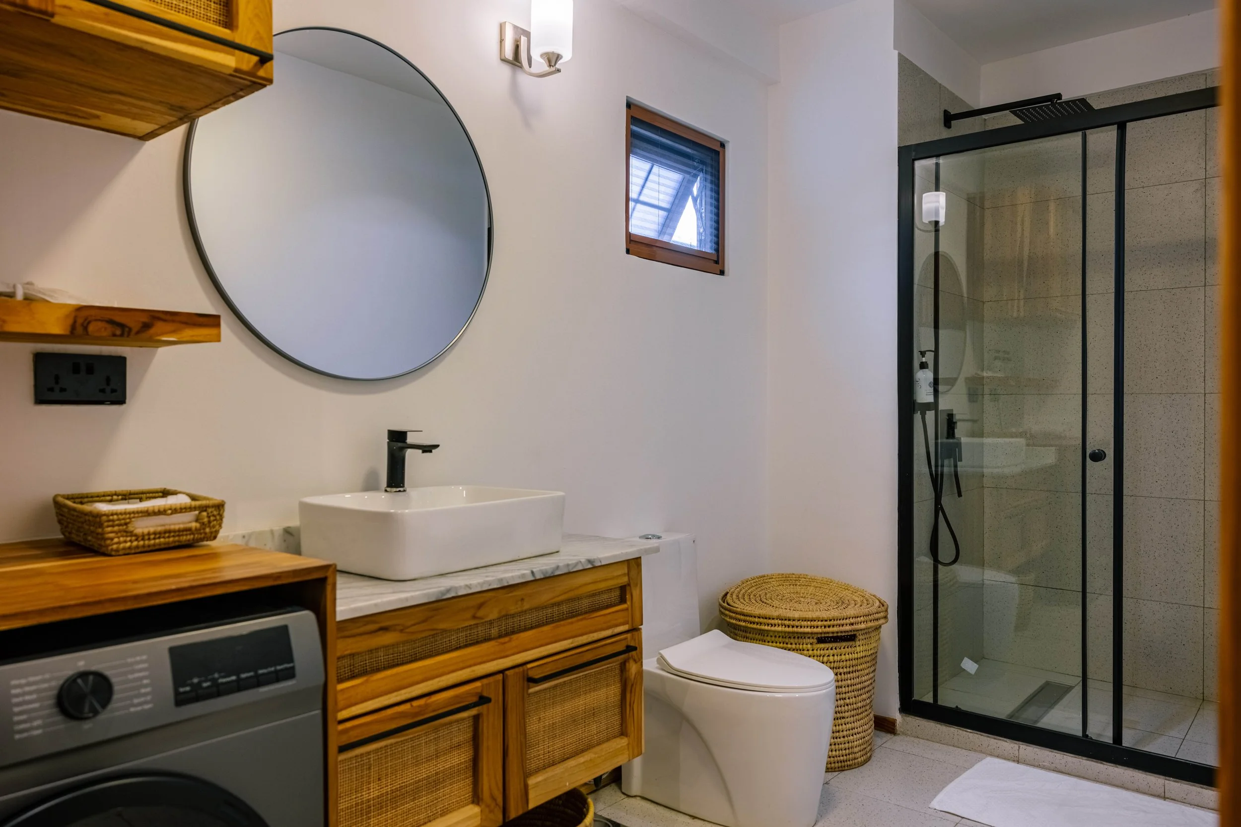 Bathroom with a wooden vanity, a round mirror, a sink, a small window, a toilet, a laundry machine, a wicker basket, a glass shower enclosure, and a white rug.