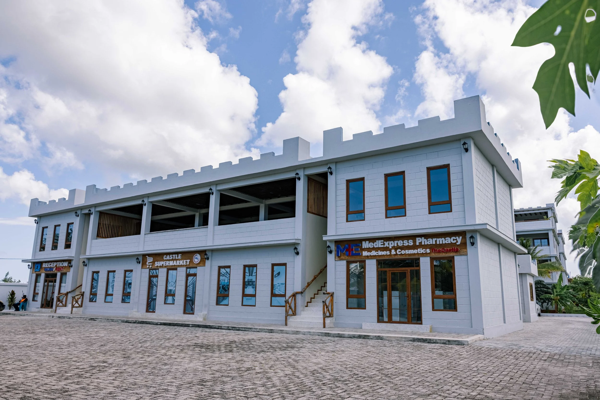 A white building with a medieval castle design features multiple businesses including a pharmacy and supermarket, under a partly cloudy sky. There are some trees and a paved area in front.