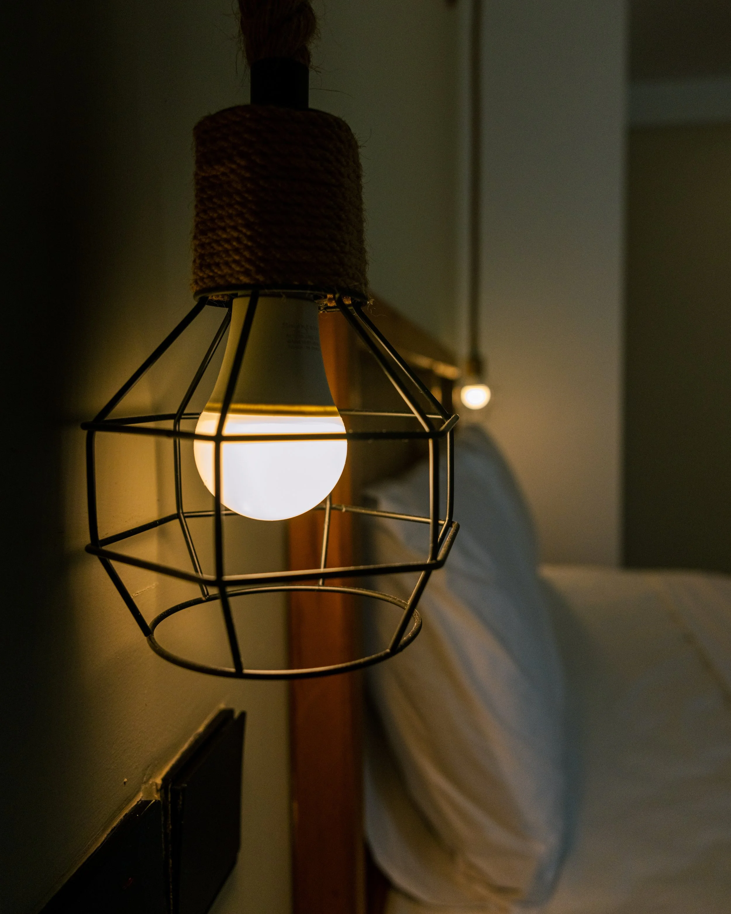 Close-up of a lightbulb inside a black metal cage on a wall, with a source of warm light illuminating the surrounding area. The fixture has a black top part with a wrapped rope or twine around it.