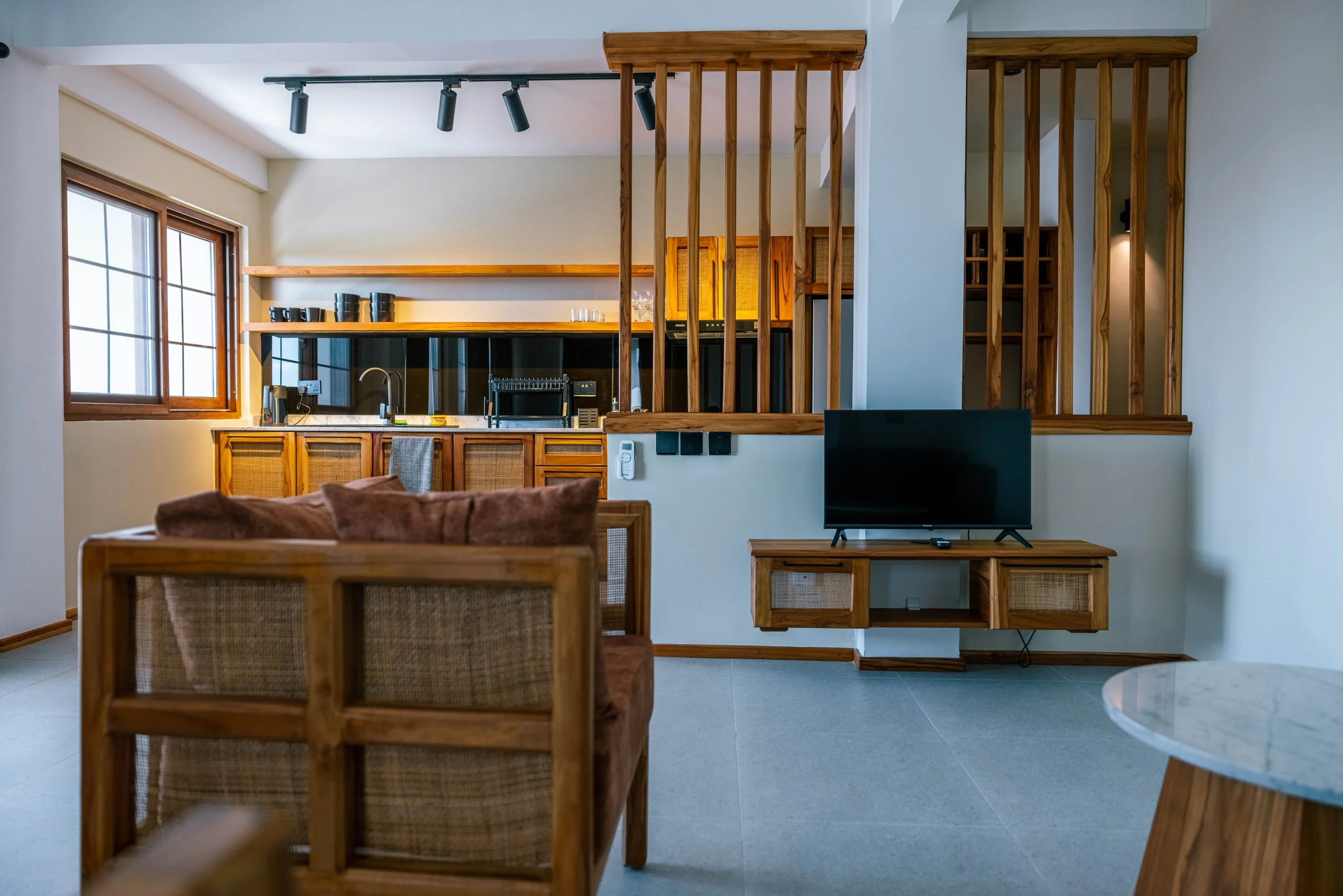 Living room with wooden furniture, including a TV stand, shelving, and a dining table with chairs, next to a kitchen area with wooden cabinets and black wall accents, illuminated by natural light from windows.